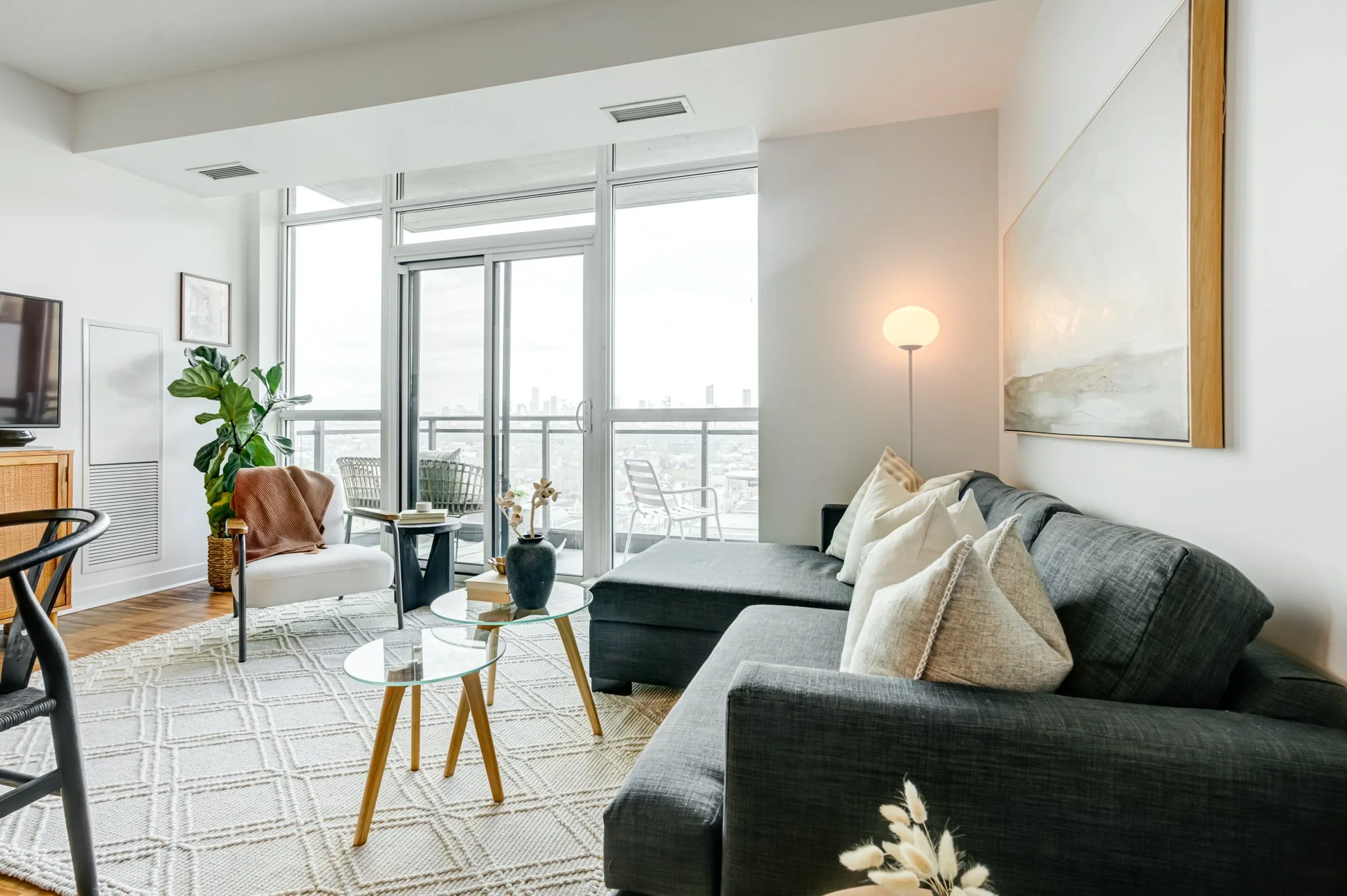 Bright living room with large windows, gray sectional sofa with pillows, glass coffee tables with a black vase and books, white and brown artwork on the wall, a floor lamp, and balcony chairs outside, with a city view.