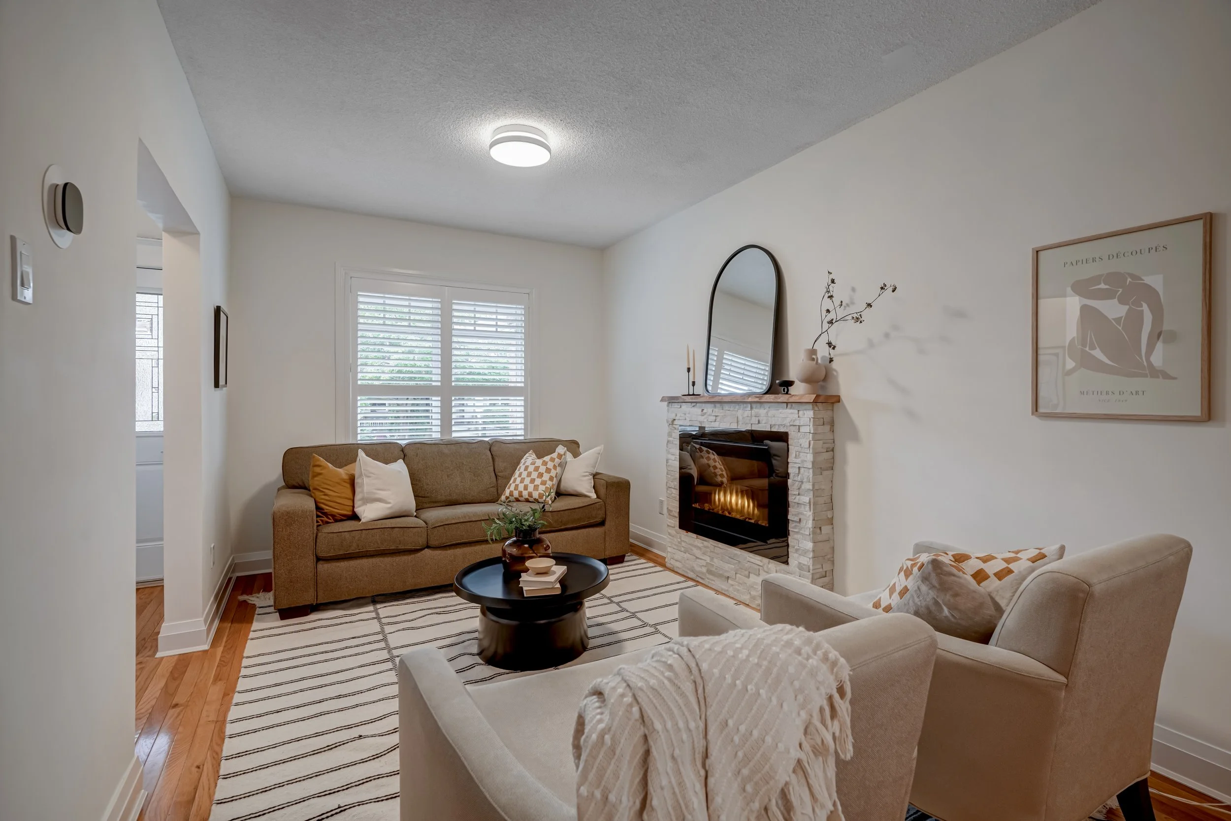 Living room with beige sofa, white armchair, fireplace with mirror, wooden mantel, window with blinds, wooden floor, striped rug, and wall art.