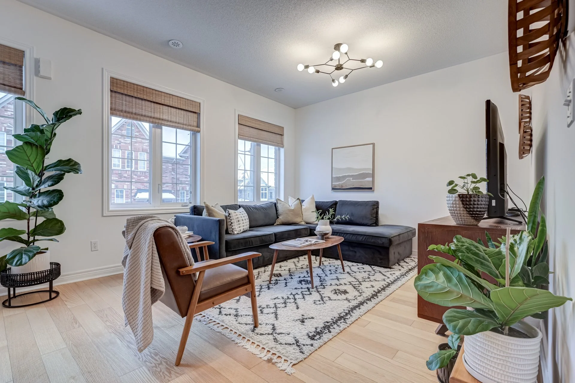 Living room with a dark gray sectional sofa, wooden coffee table, beige armchair, potted plants, artwork on white walls, windows with bamboo blinds, and a beige patterned rug.