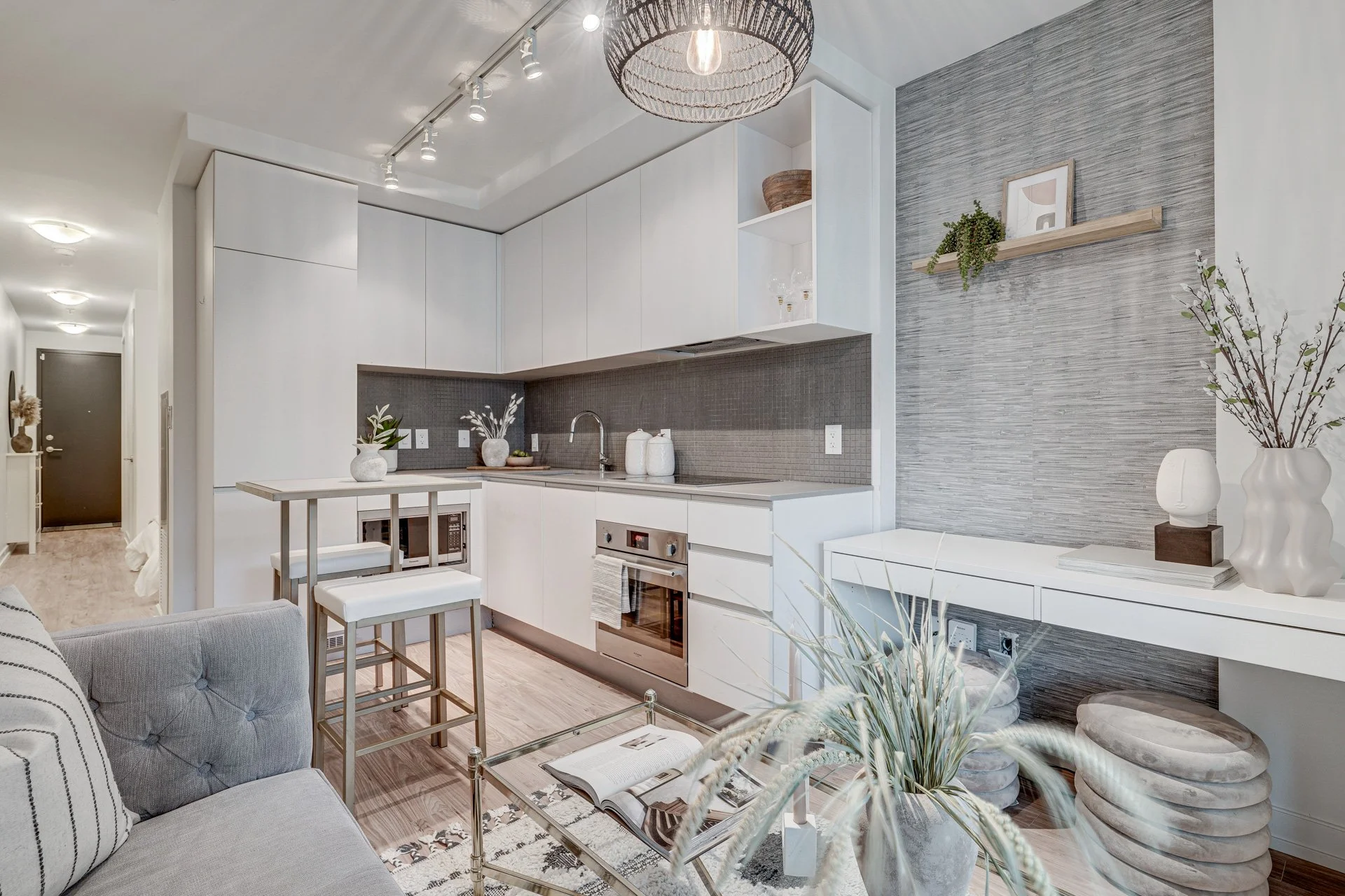 Modern kitchen and living room with light gray and white decor, including a gray sofa, glass coffee table, white cabinets, gray backsplash, and decorative plants and picture frames.