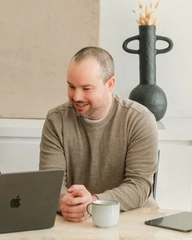 A man with a shaved head and beard smiling while working on a laptop at a table, with a white mug and a closed tablet nearby, and a decorative black vase with dried flowers in the background.