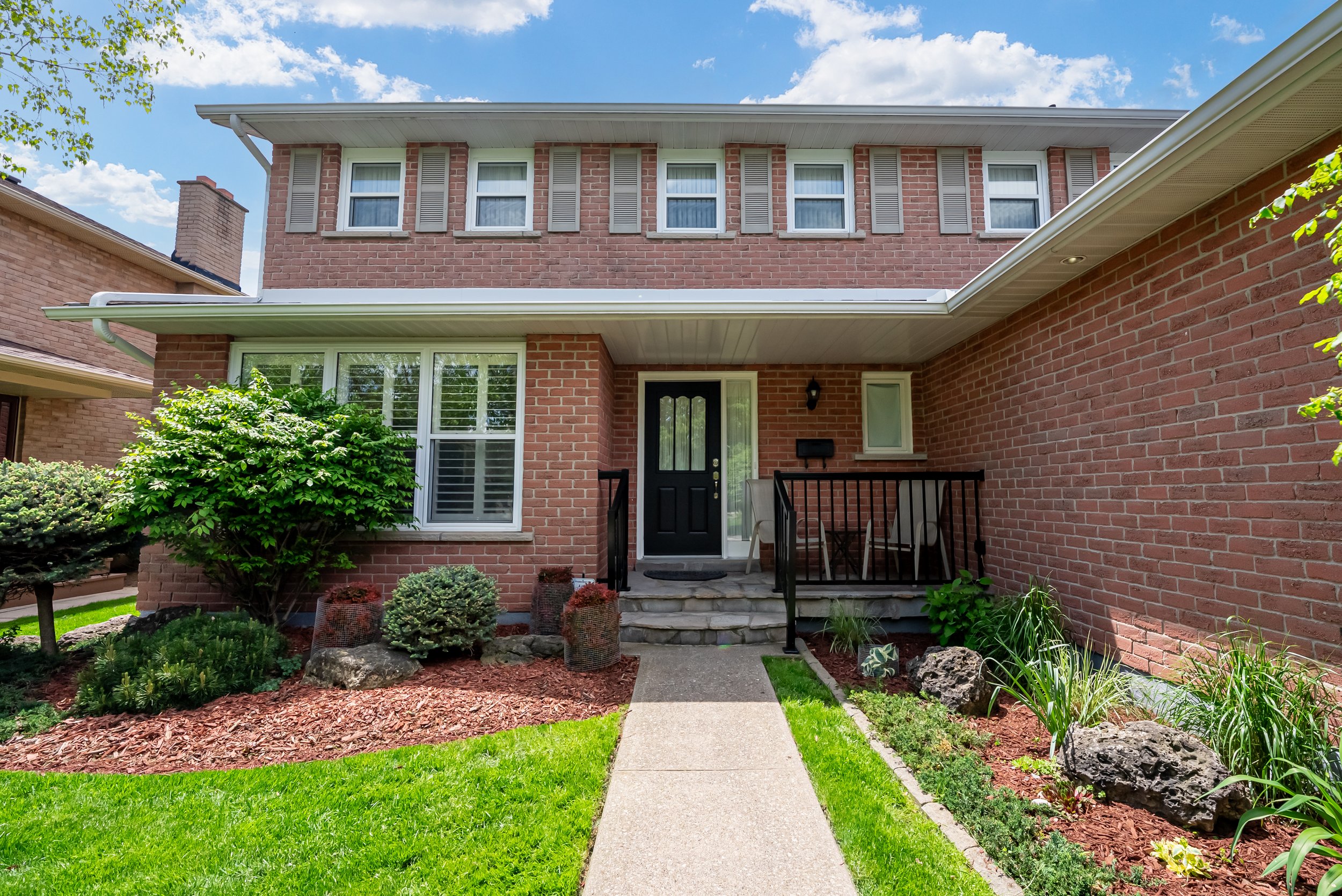 Front entrance of a brick house with a black door, small porch with steps, and landscaped garden with bushes, rocks, and flowers.