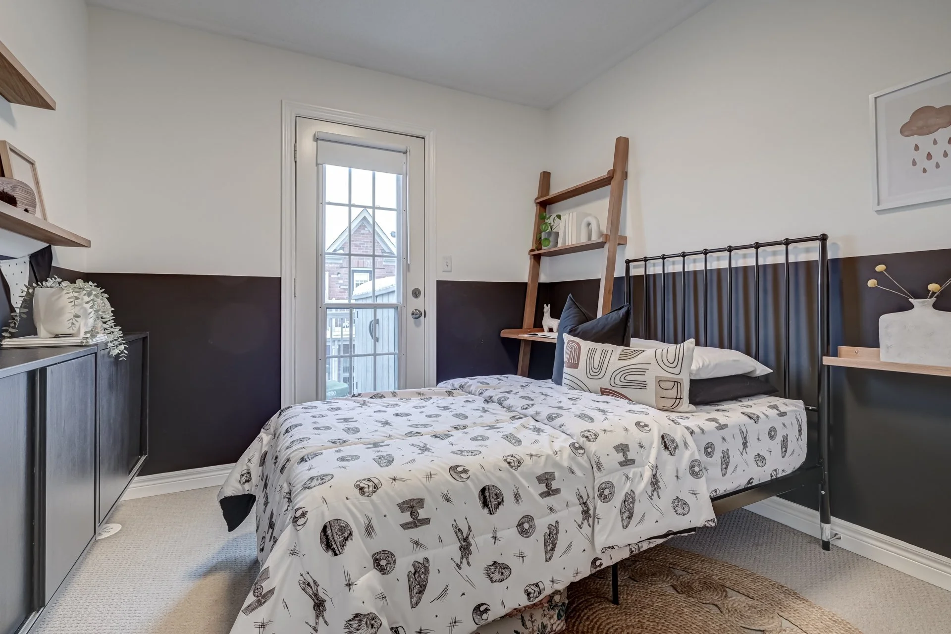 A bedroom with a bed featuring patterned bedding and decorative pillows, beside a wooden ladder-style shelf with plants and decor, a black and white wall art, a dark cabinets with decor, a glass door leading to a balcony, and a rug on a beige carpet.