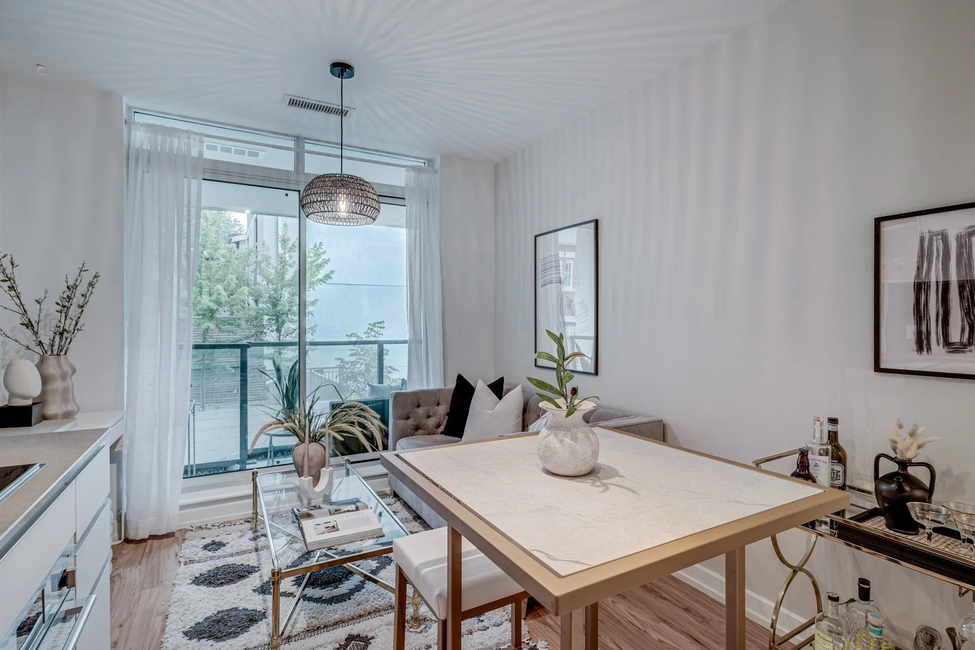 Modern living room with pastels, a large window, a gray sofa, decorative pillows, artwork on the walls, a marble dining table with a plant centerpiece, bar cart with bottles, and stylish decor.