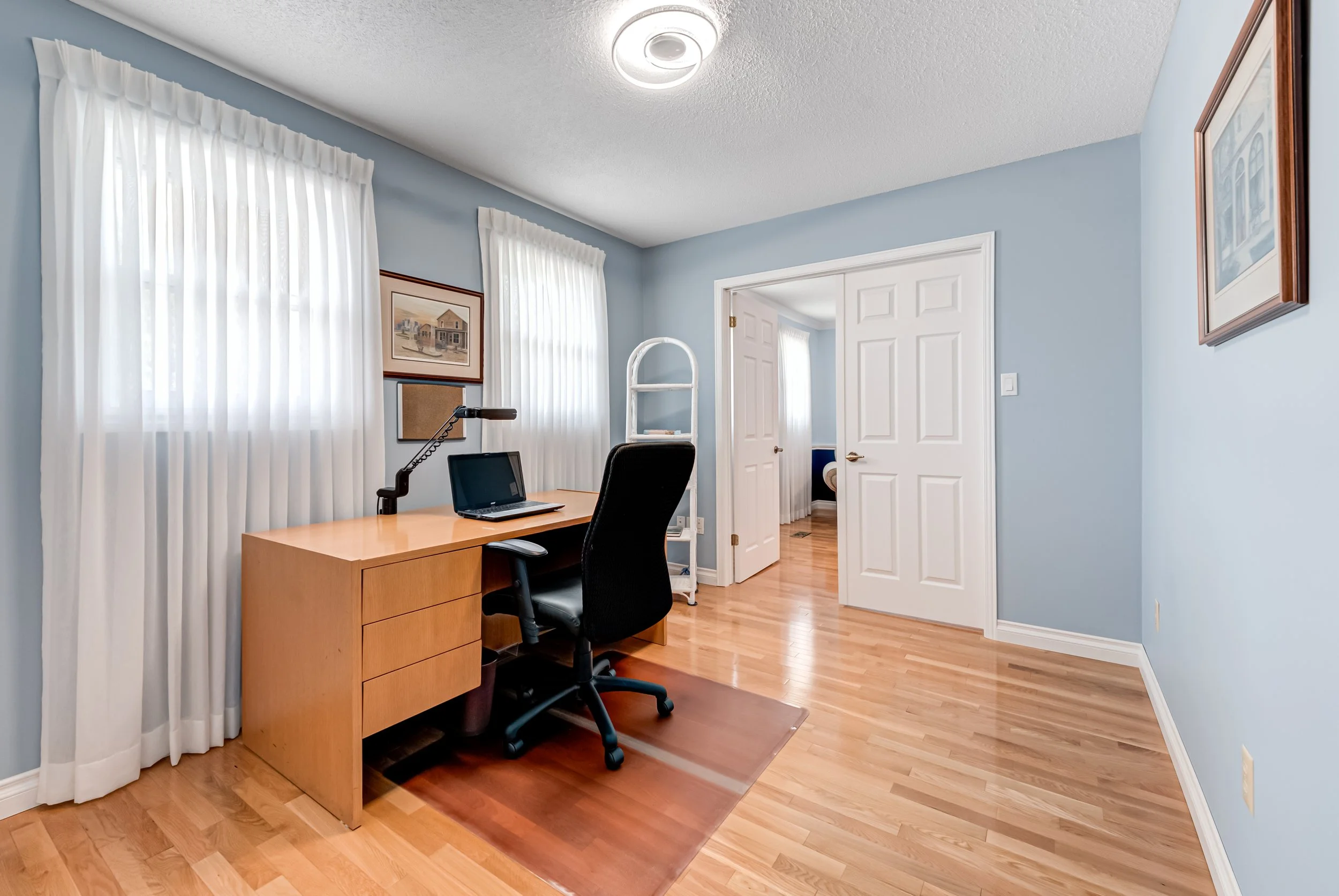 A home office with light blue walls, wooden floor, white curtains, a wooden desk with a laptop, and a black office chair.
