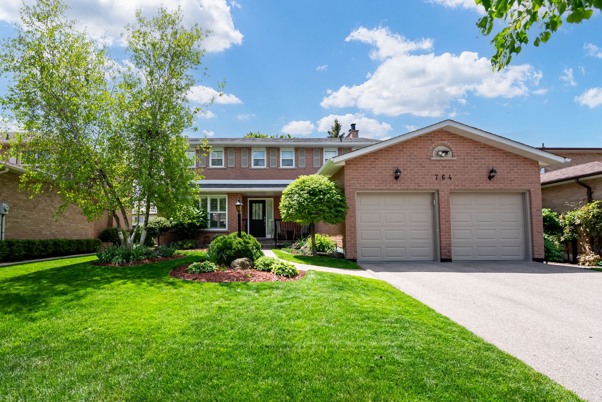 A suburban house with a brick facade, a double garage, and a well-maintained front yard with green grass, shrubs, and trees under a partly cloudy sky.