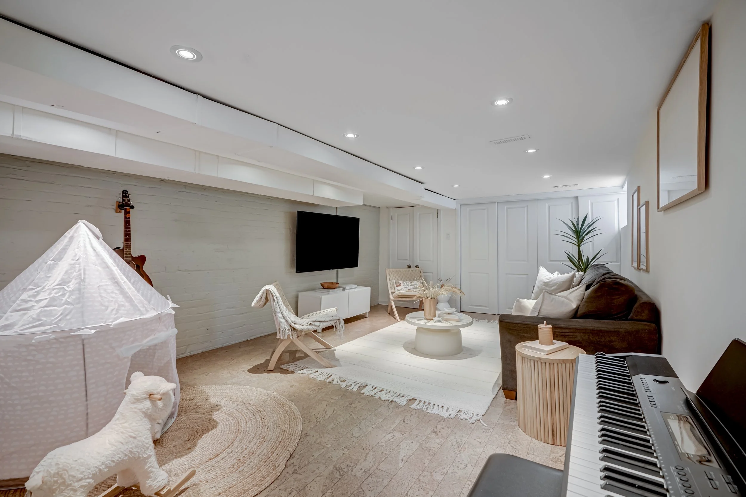 A cozy living room with white walls and ceiling, a large flat-screen TV, white brick accent wall, and various furniture including a dark sofa with white cushions. There's a round coffee table, a small side table with a candle, a keyboard, and a tent 