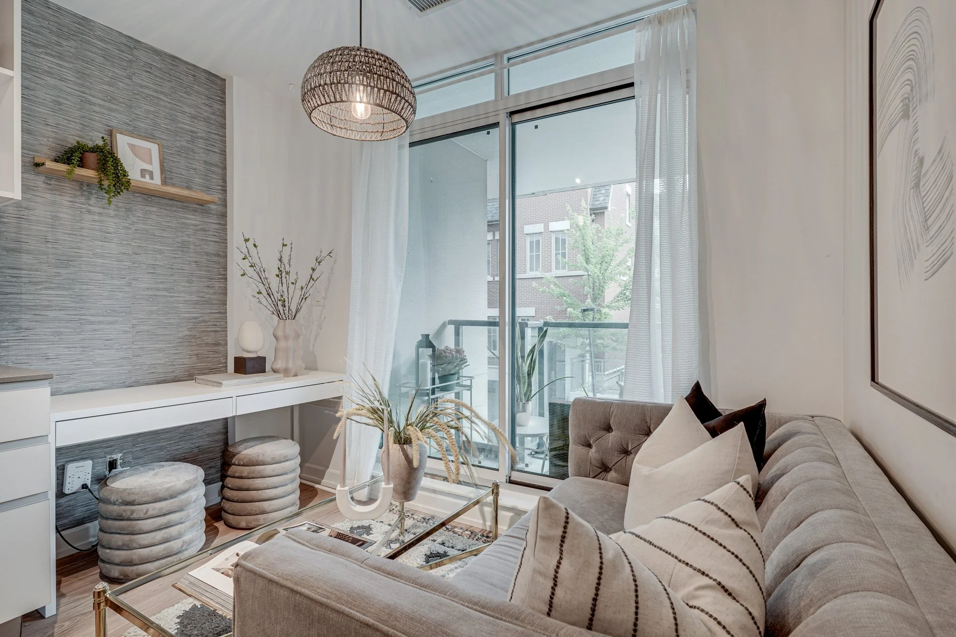 Living room with a light gray tufted sofa, decorative pillows, a glass coffee table with plants, a large sliding glass door leading to a balcony, sheer white curtains, a textured gray accent wall with a floating shelf, a wicker ceiling light, and var
