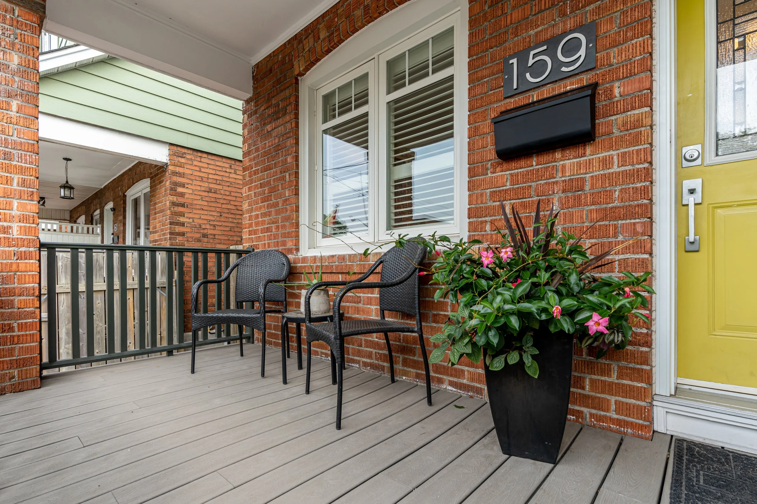 Front porch with two black chairs, a large potted pink flower plant, brick walls, yellow door, and house number 159.