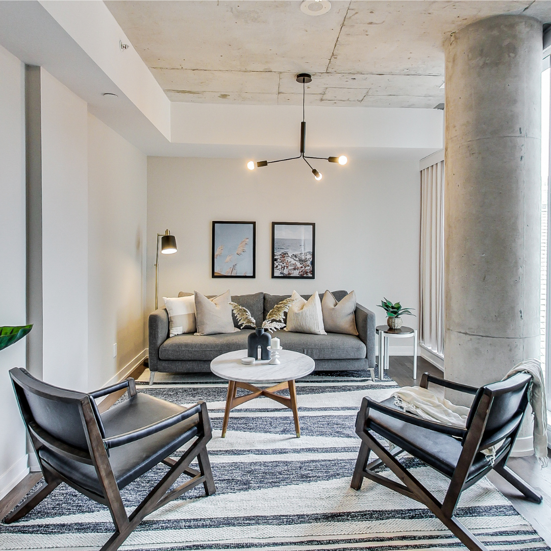 Modern living room with a gray sofa, two black armchairs, a round marble coffee table, a striped area rug, a concrete ceiling, a large concrete pillar, and decorative wall art.