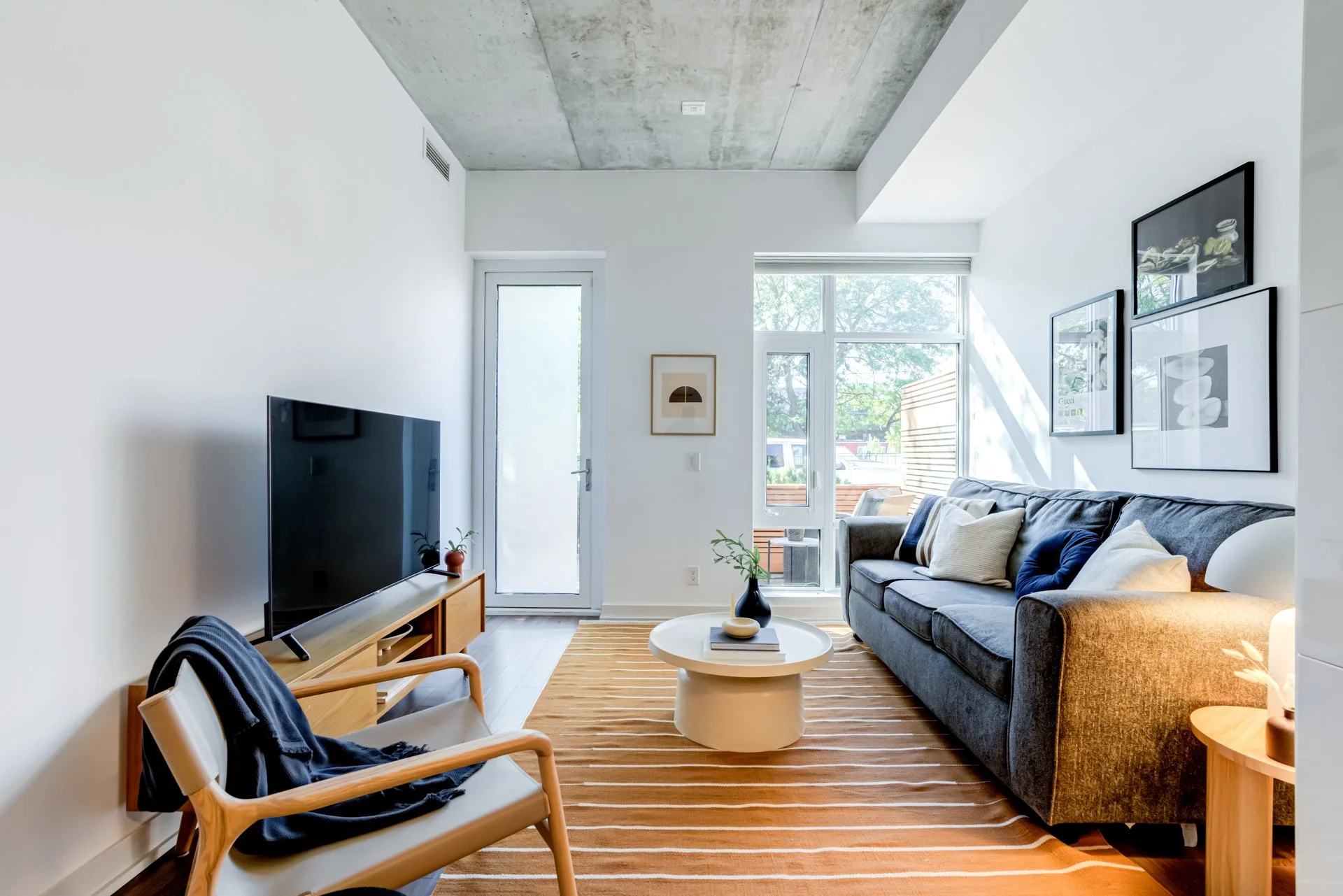 Modern living room with a gray sofa, wooden side table, and white coffee table on an orange striped rug, with a large window, abstract artwork, a flat-screen TV on a wooden stand, and a cozy chair with a blanket.