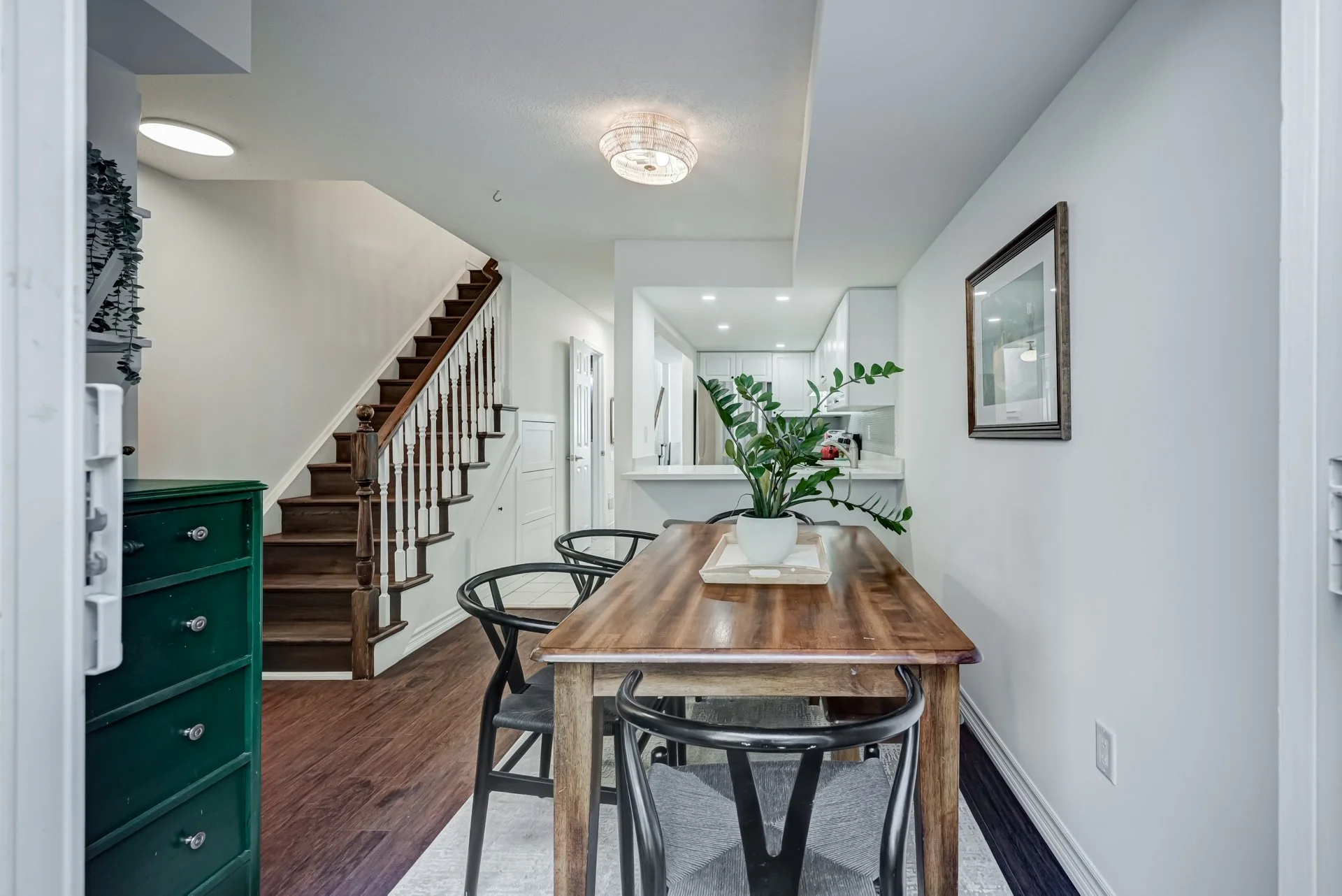Dining area with wooden table, black chairs, and a green potted plant, adjacent to a staircase in a bright, modern home.