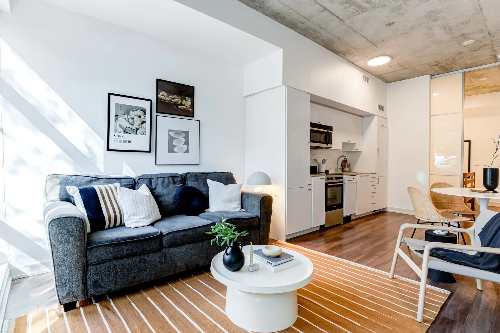 Modern living room with a gray sofa, white coffee table, and a small plant. Adjacent to an open kitchen with white cabinets and wooden flooring. Dining area with a round table and woven chairs, in a space with minimalist decor.