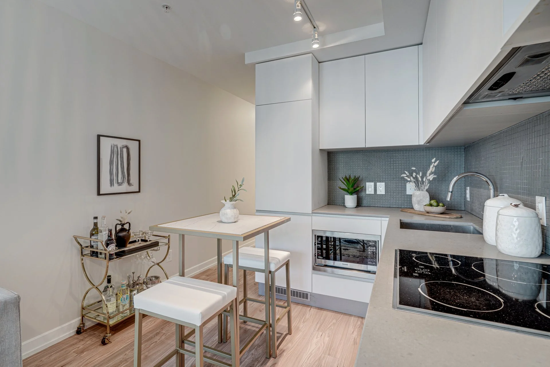 Modern kitchen with white cabinets, gray backsplash, and wooden floor. There is a small dining area with two white stools, a bar cart with bottles, and decorative vases and plants on the counter and wall.
