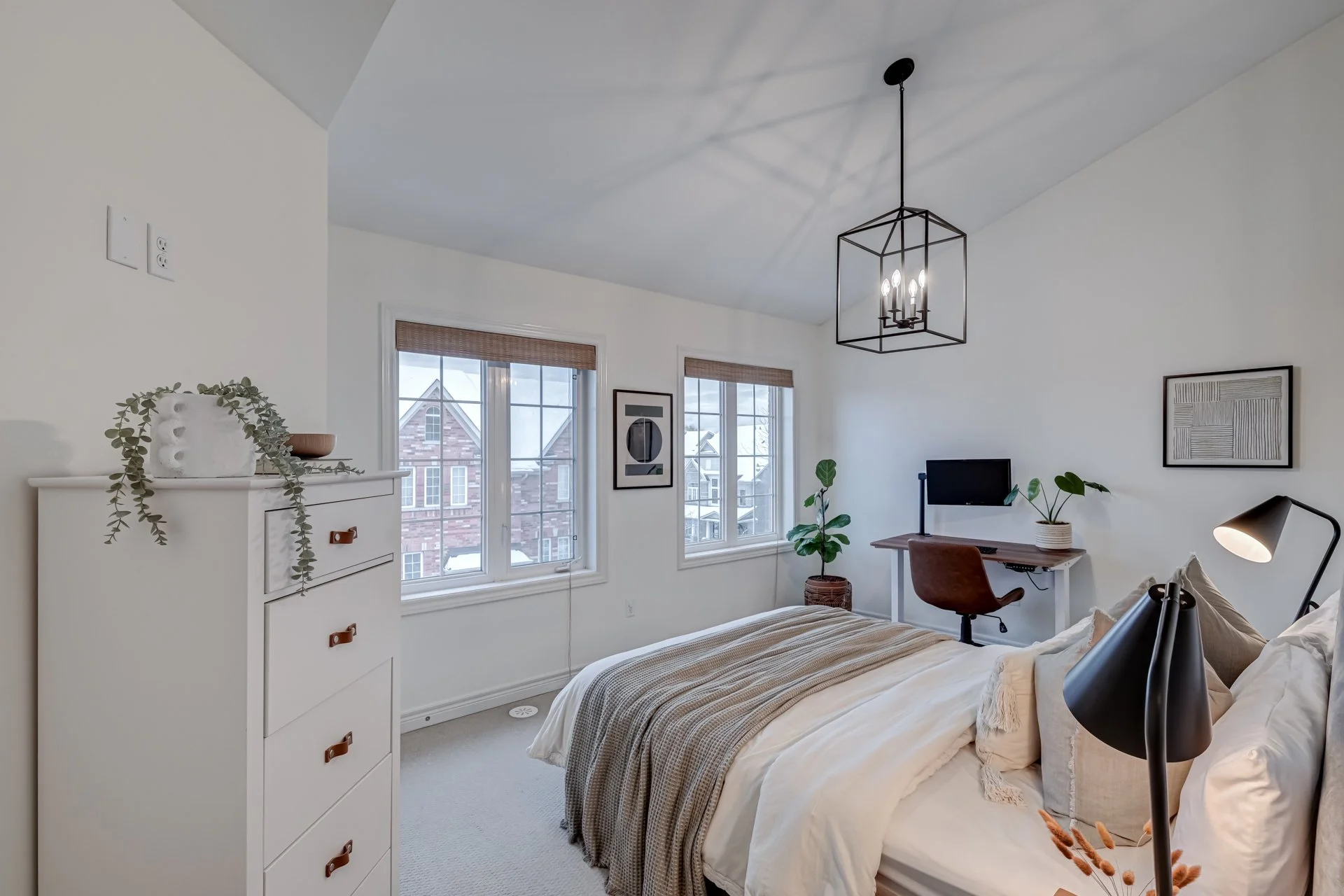 A bedroom with white walls, a bed with beige and white bedding, a white dresser with decor, two windows with wooden blinds, and a small desk with a computer and plants. A black geometric chandelier hangs from the ceiling.