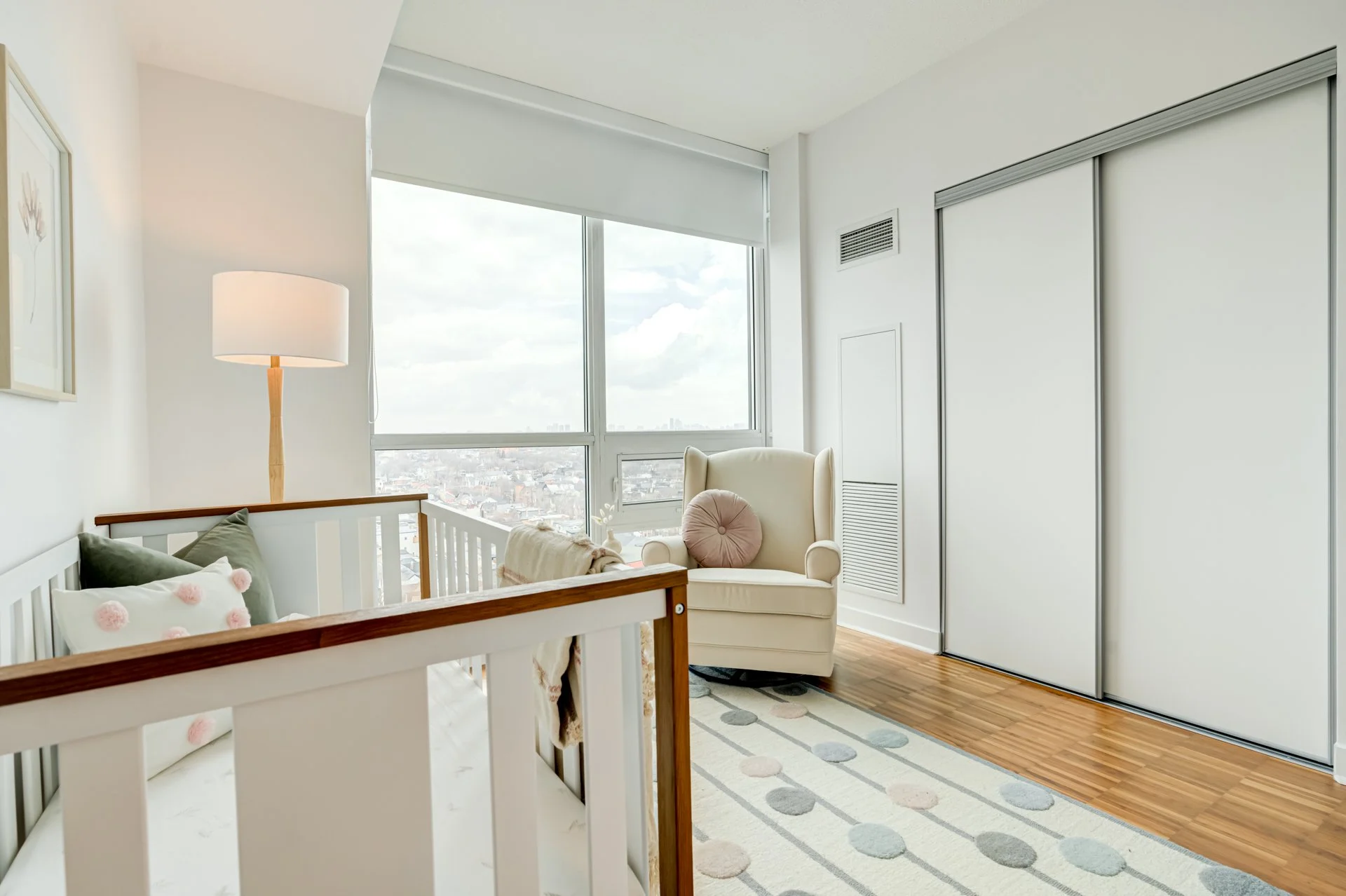 Bright nursery with white walls, a crib with pillows, a plush armchair by large windows, a floor lamp, and a colorful polka dot rug.