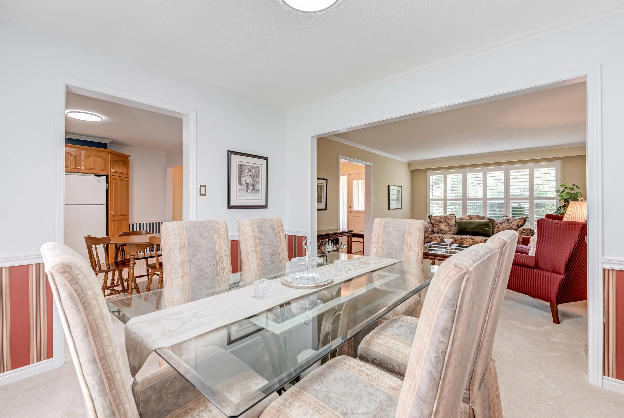 A dining room with a glass top table surrounded by eight upholstered chairs. In the background, a living room with large windows, a red armchair, a floral sofa, and a small table with a lamp. An open doorway reveals a kitchen with wooden cabinetry an