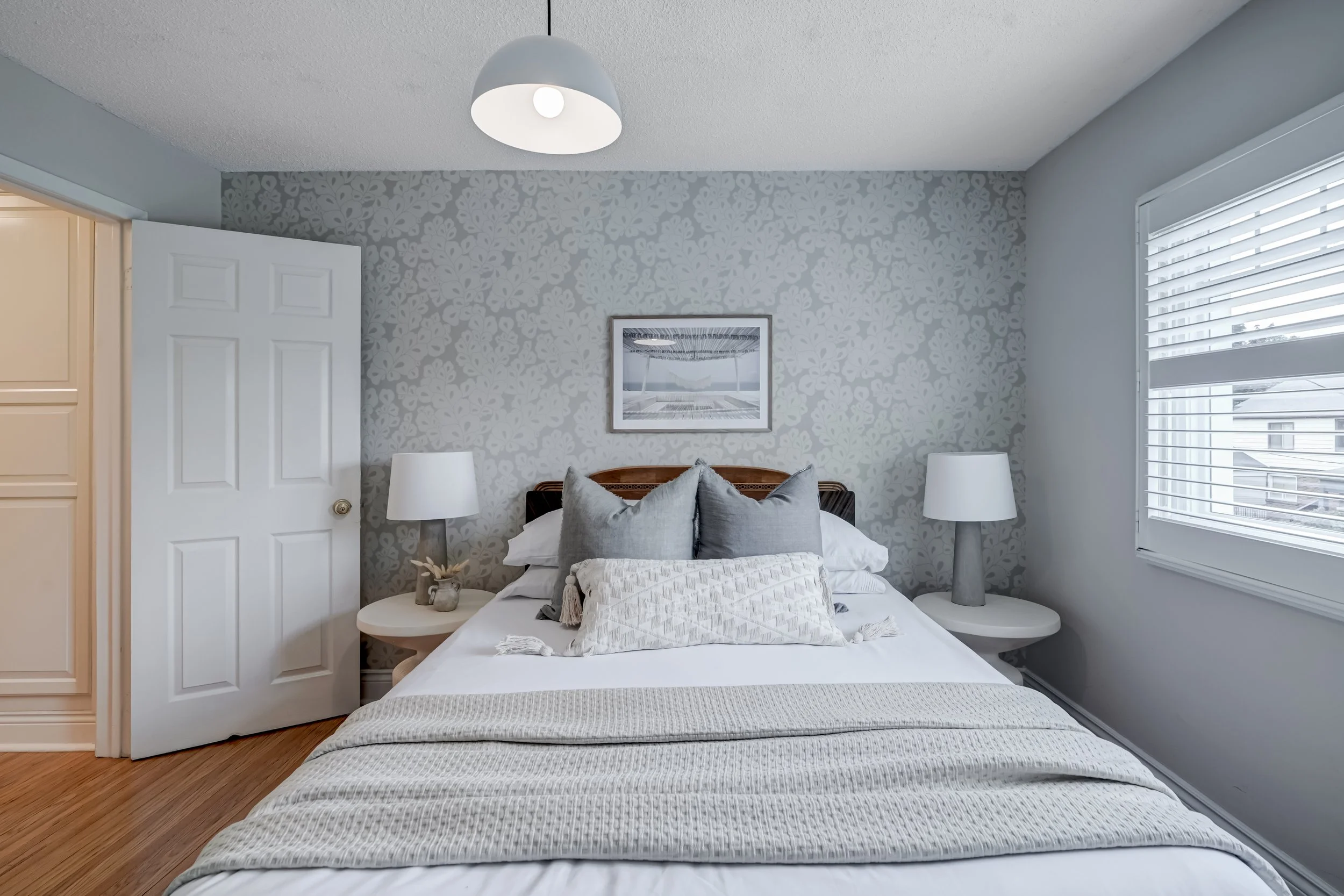 A neatly made bed with gray pillows, a cream-colored throw blanket, and a decorative pillow in a bedroom with light gray and floral patterned wallpaper, two matching nightstands with lamps, a window with white blinds, and a door slightly ajar.