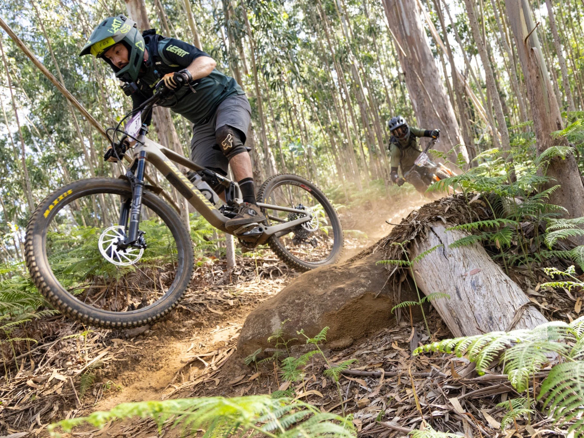 Two mountain bikers racing on a dirt trail through a forest, with one leading and the other following close behind, kicking up dust as they navigate the rugged terrain.