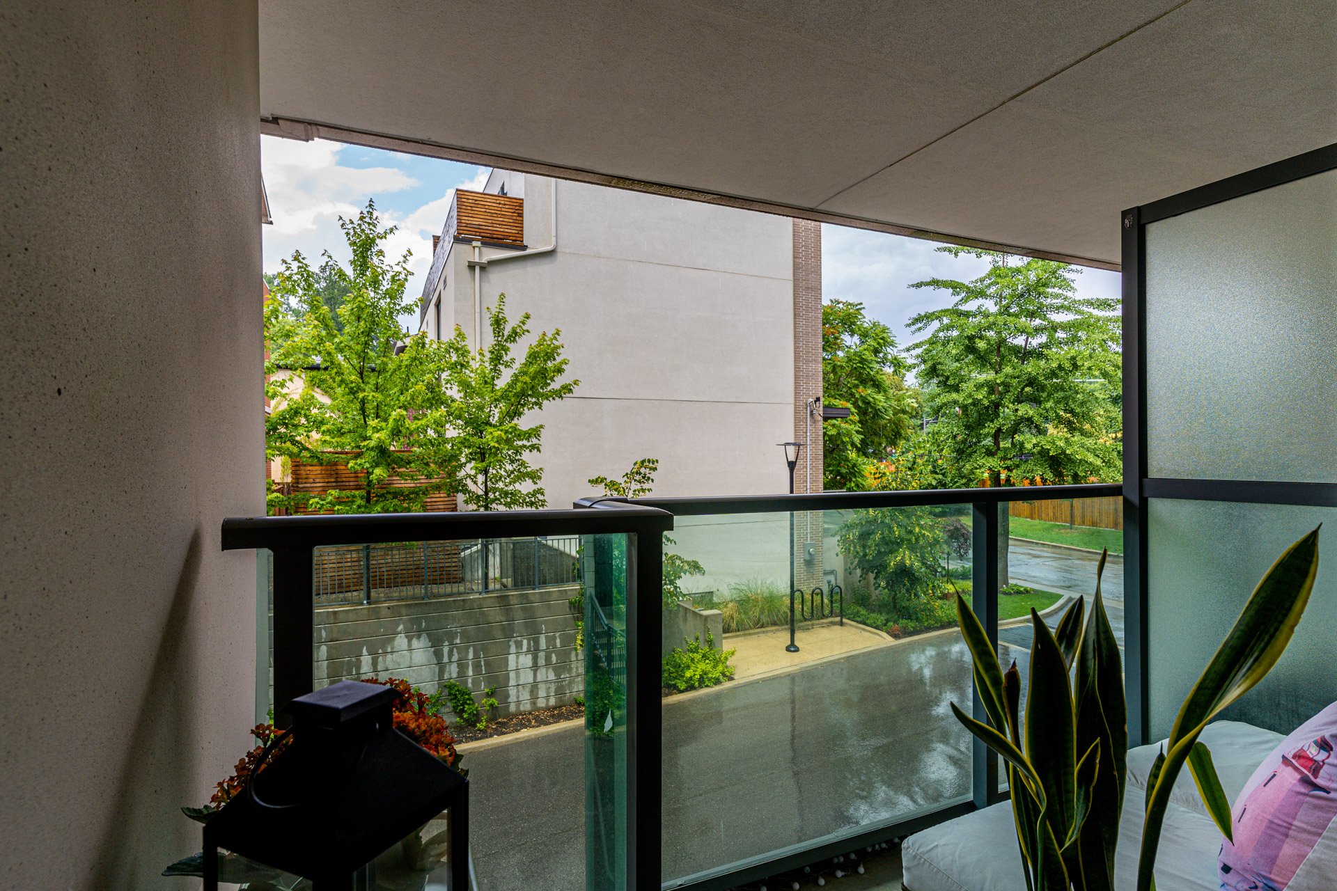 View from a balcony showing green trees, a sidewalk, and apartment buildings in the background on a rainy day.