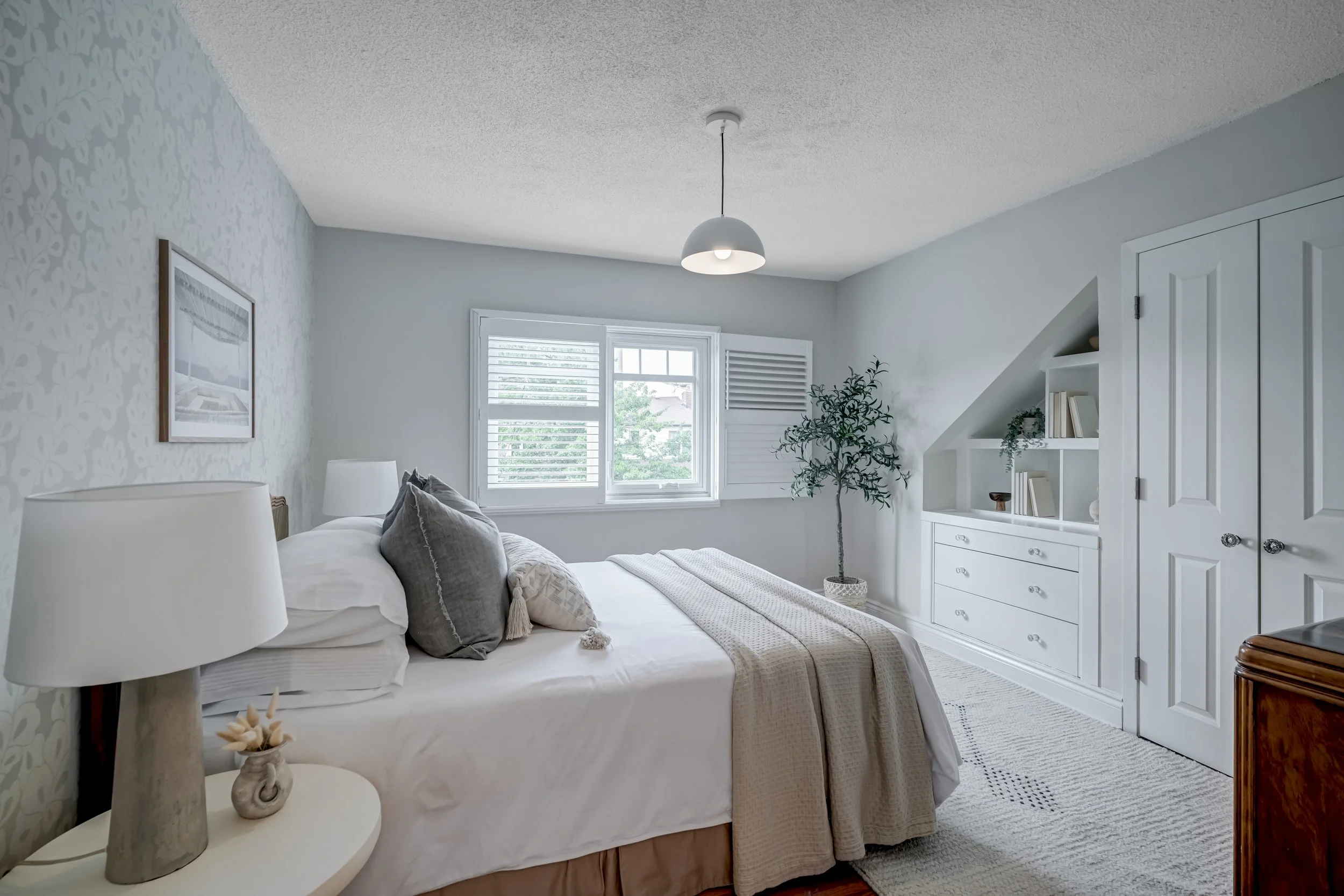 A bedroom with light-colored walls, a bed with white bedding and gray pillows, a nightstand with a lamp and decorative item, a window with blinds, a potted plant, built-in shelves with books and decor, and a closed white door.