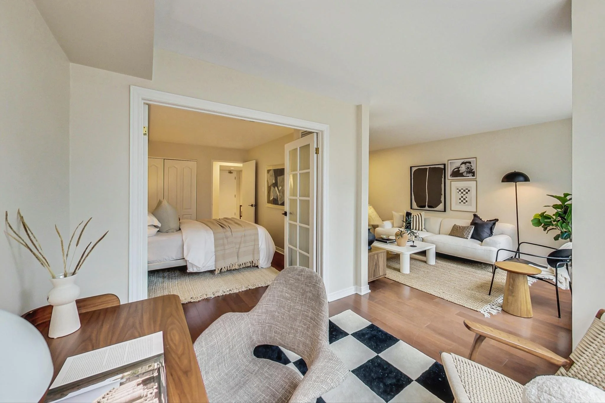 Interior of a modern apartment with a bedroom and a living room, featuring neutral-toned furniture, wooden flooring, and decorative wall art.