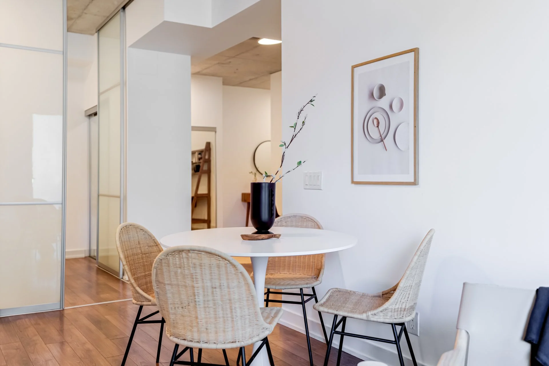 A white round dining table with a black vase containing branches in a modern, minimalist room with wooden flooring and white walls.
