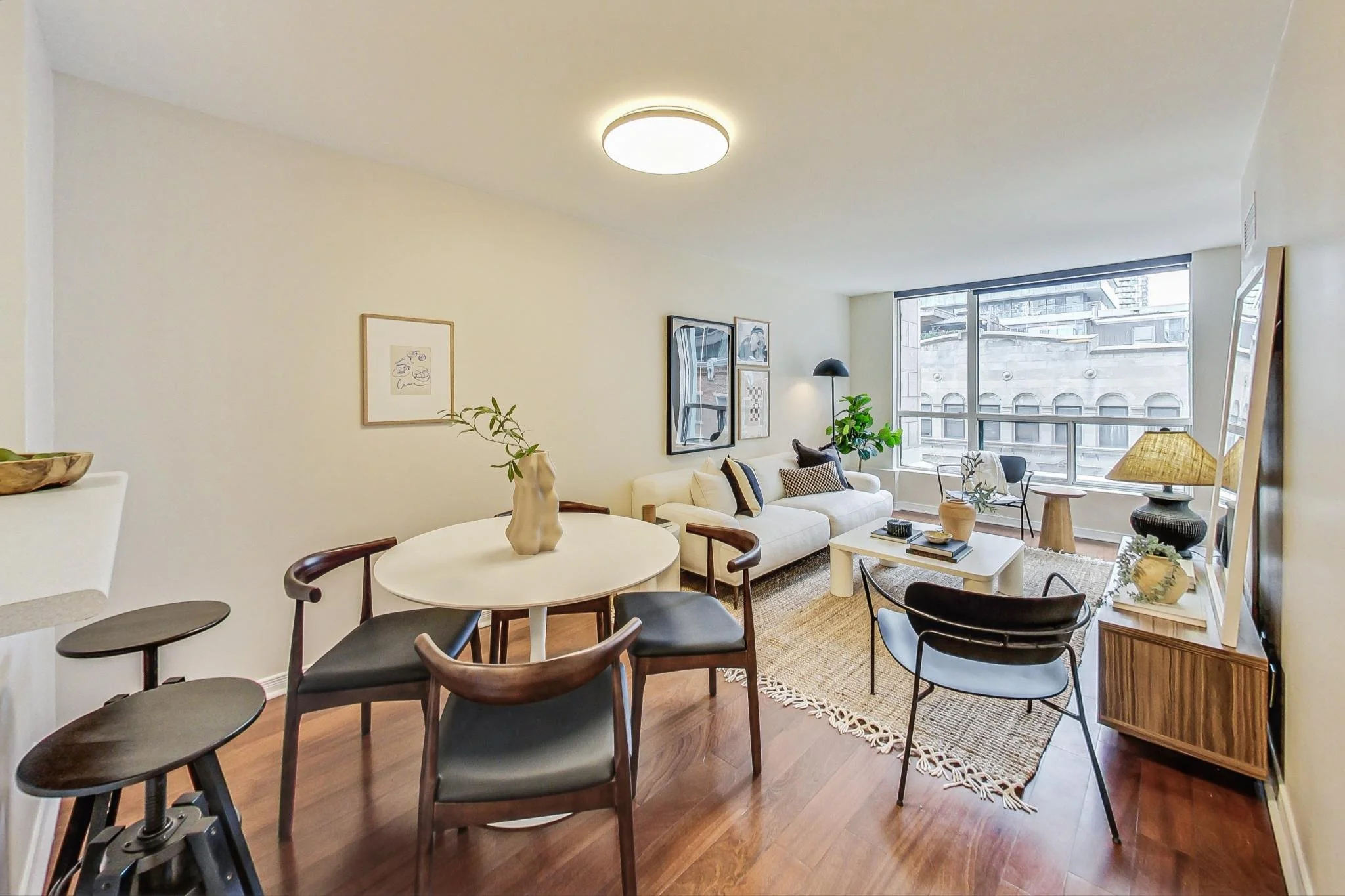 Open living room and dining area in an apartment with white walls, hardwood floors, a white couch with cushions, a round dining table with chairs, artwork on the walls, a large window, and various decorative items.
