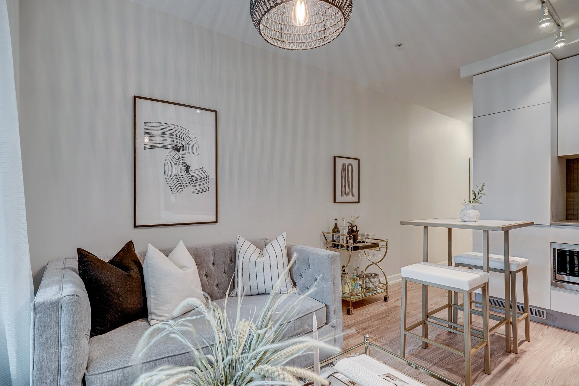 Modern living room with light-colored sofa, decorative pillows, wall art, bar cart, and a small table with a vase and a plant.