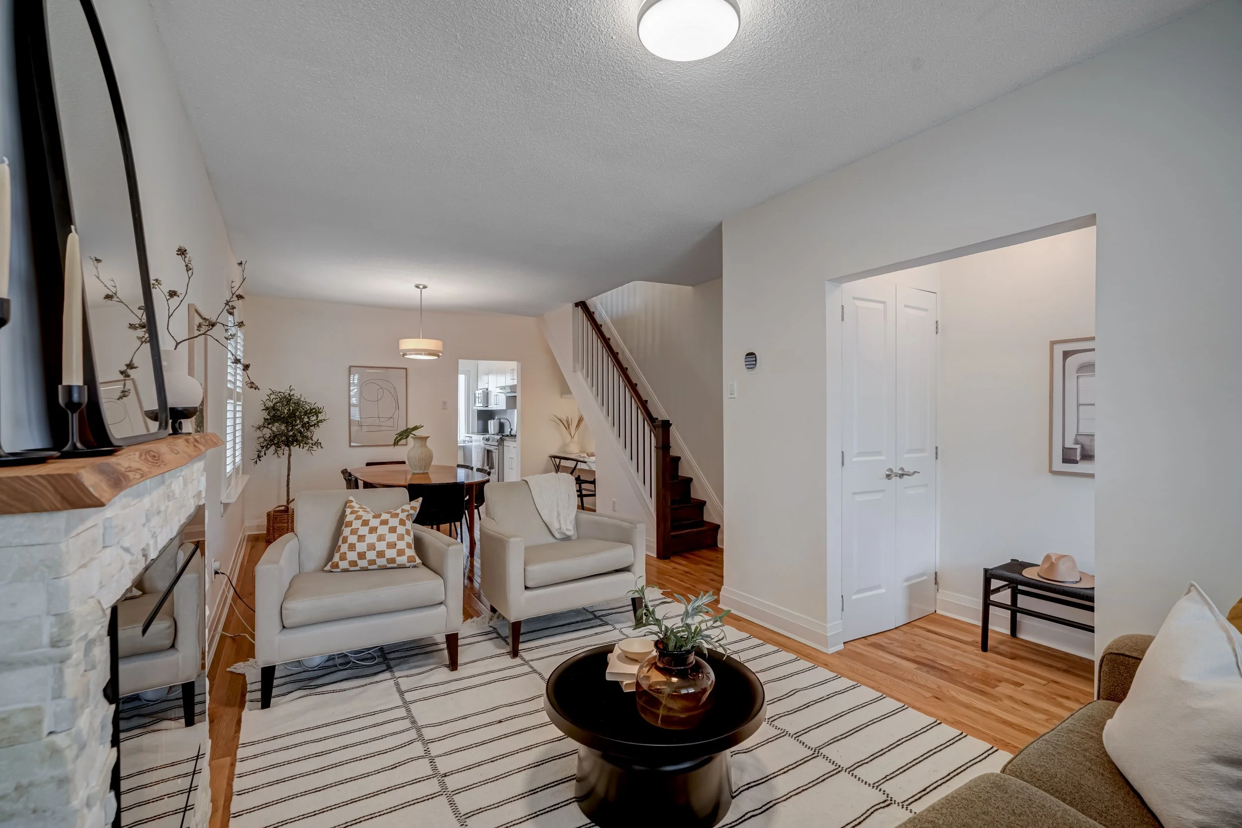 Living room with white walls, hardwood floor, and furniture including two white armchairs, a coffee table with a plant, and a beige sofa. Visible staircase, dining area with round table and chairs, and a kitchen in the background.