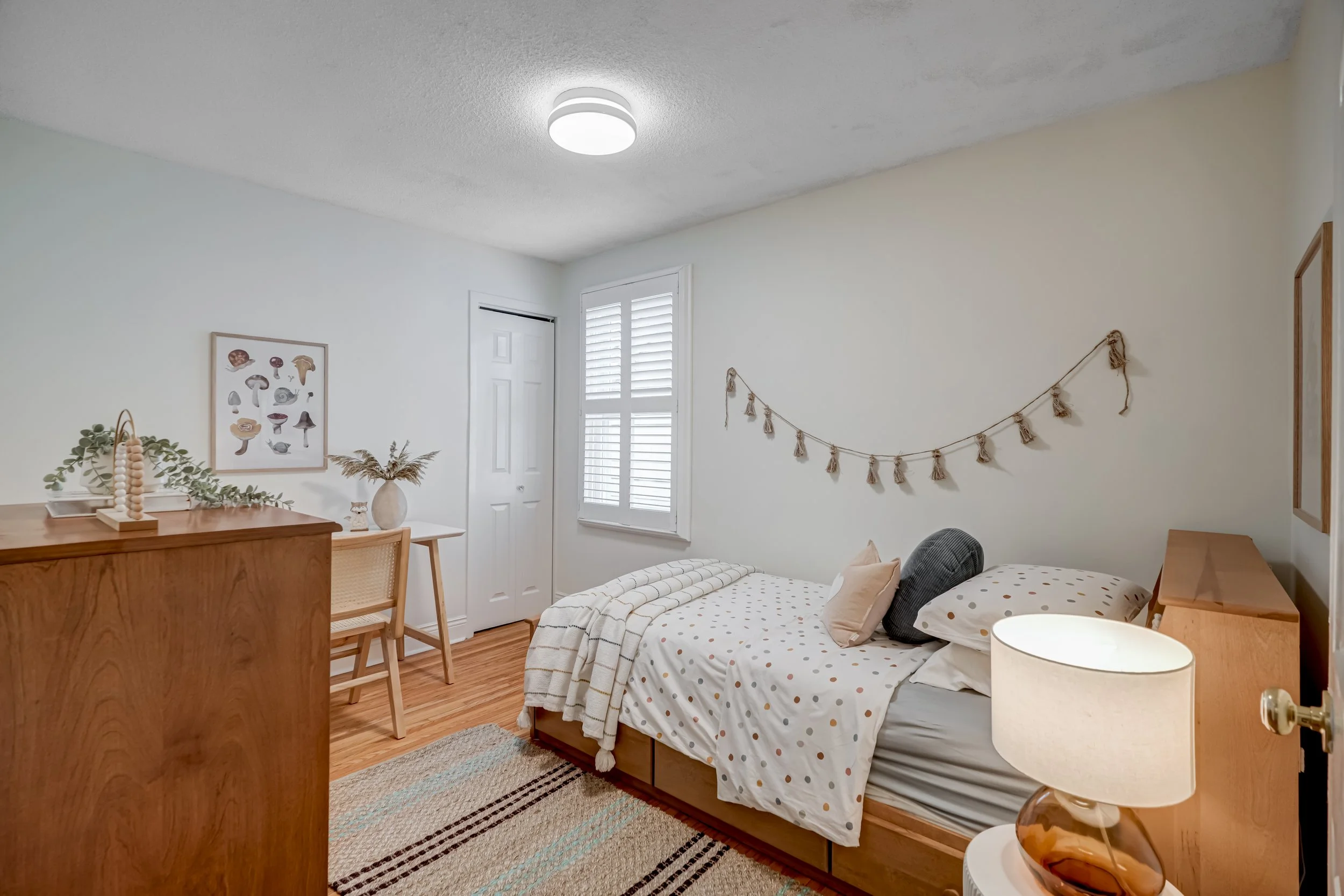 A cozy bedroom with a bed covered in a white polka dot duvet, decorated with pillows, a beige throw, and a garland on the wall. There's a window with white shutters, a wooden dresser, a small desk with a chair, a lamp, and artwork featuring mushrooms