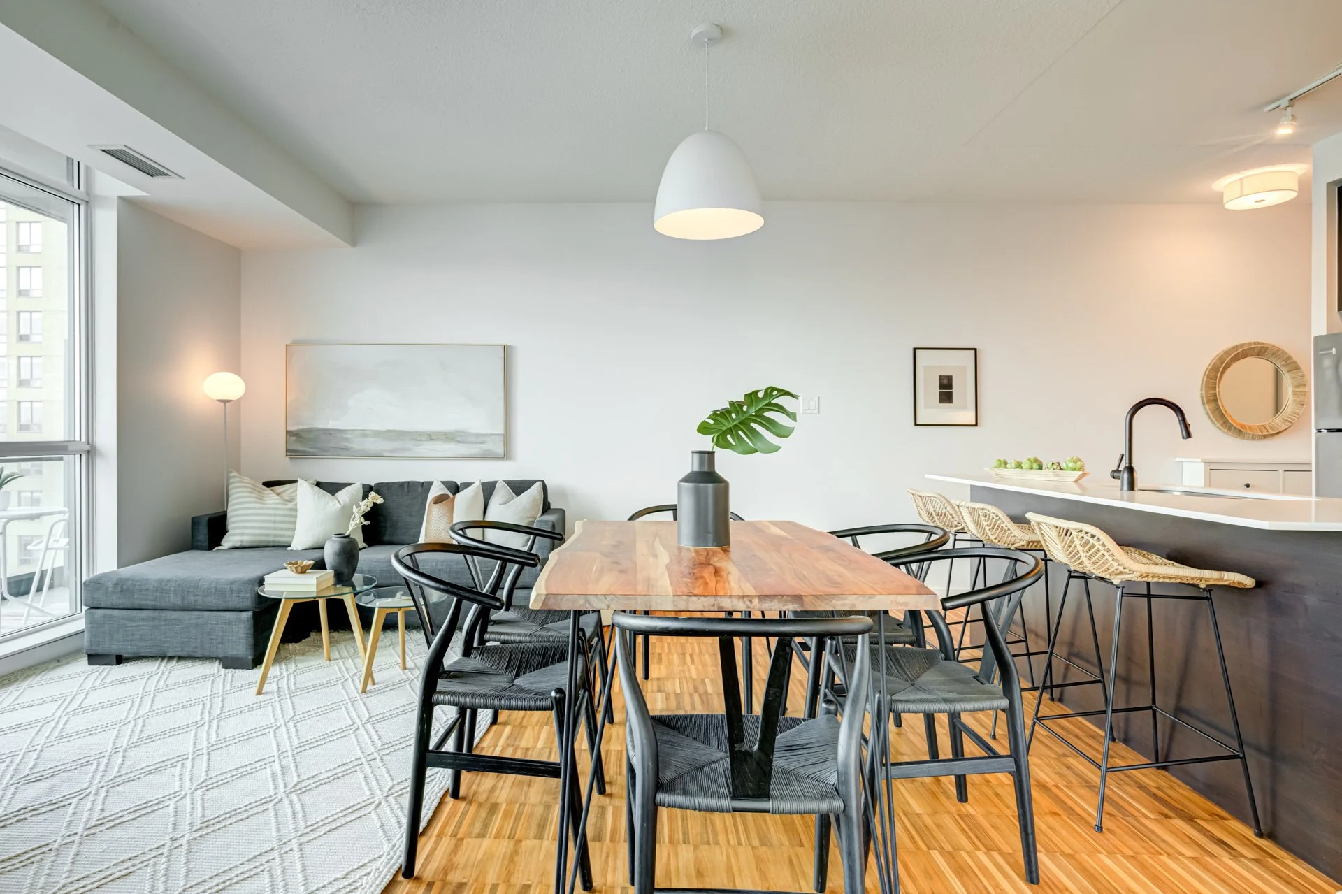 Open-concept living and dining area with a wooden dining table and six black chairs, a grey sofa with multiple cushions, a small side table, and a modern kitchen with bar stools and a mirror on the wall. Bright natural light from large windows.