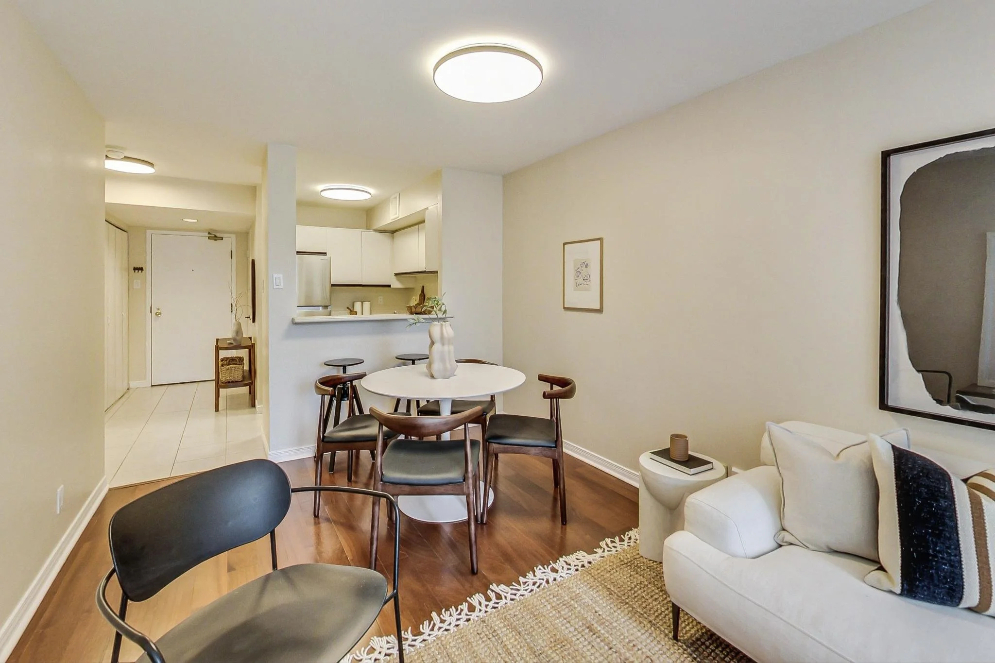 Living room with white sofa, black armchair, and wooden dining table with six chairs; kitchen in background with white cabinets and stainless steel appliances.