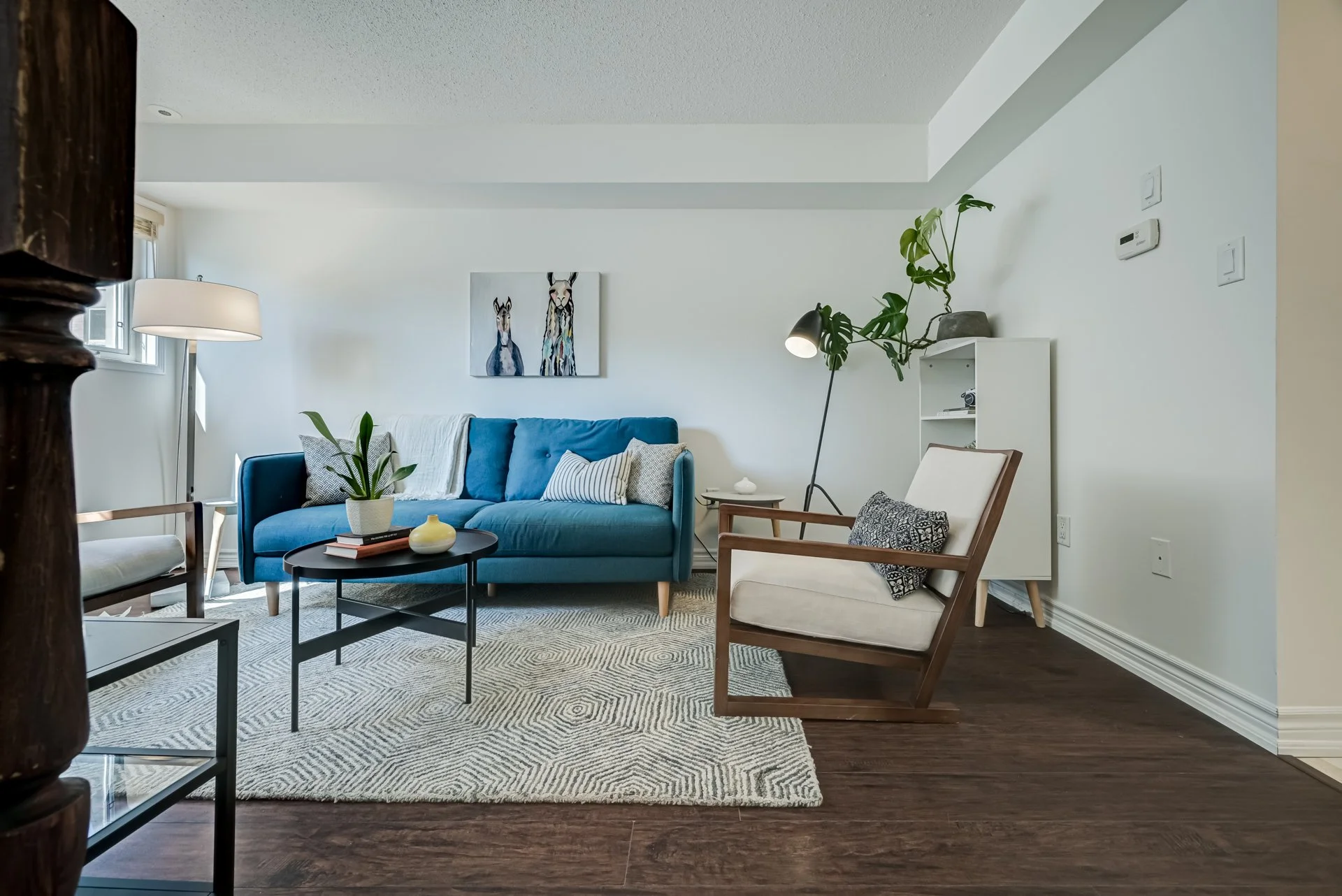 Living room with a blue sofa, white armchair, modern black coffee table, and a white shelving unit. Decor includes artwork of two animals hanging on the wall, potted plants, and a patterned rug on dark wood flooring.