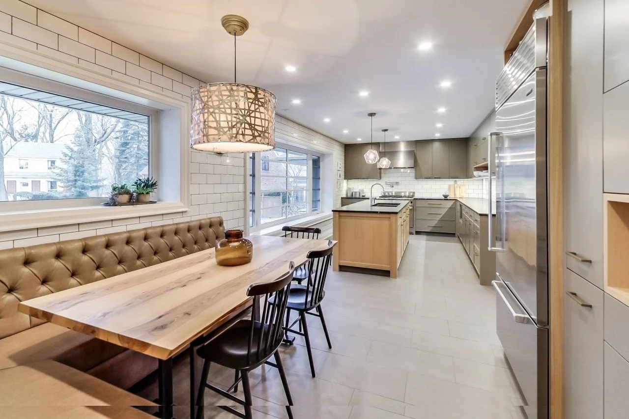 Modern kitchen with white brick walls, large windows with a wintery outdoor scene, a dining area with a wooden table and black chairs, and a spacious counter with gray cabinets and stainless steel appliances.