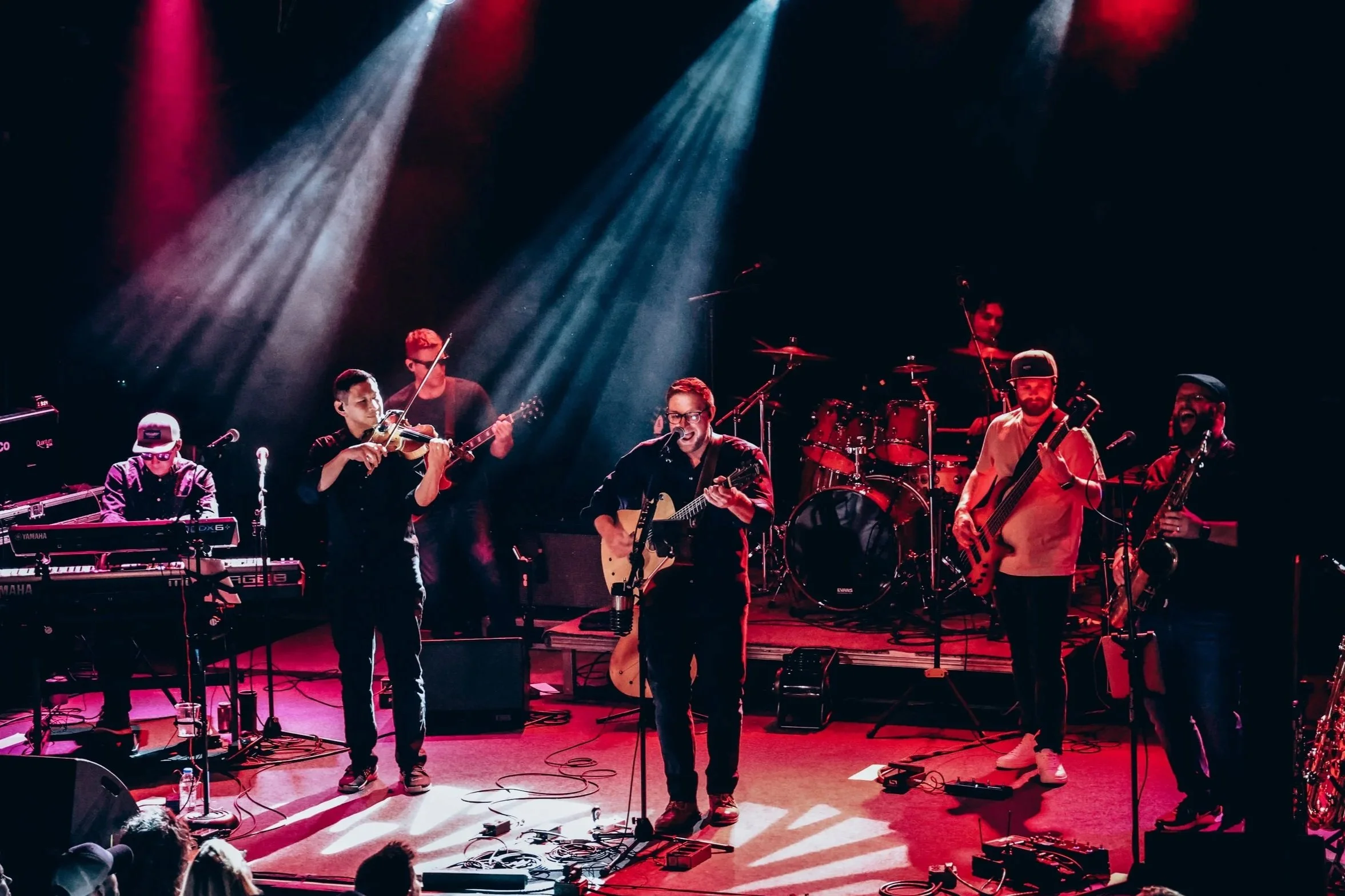 Band performing on stage with multiple musicians playing instruments under colorful stage lighting