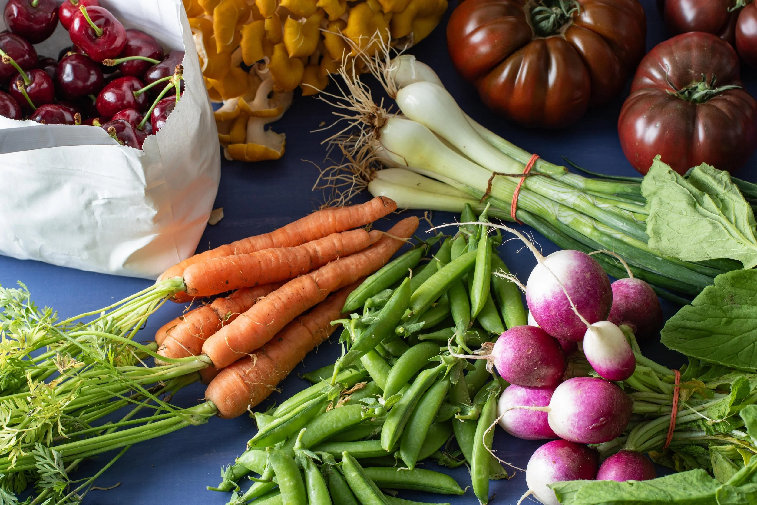Various fresh vegetables including tomatoes, carrots, radishes, green beans, green onions, cherries, and yellow mushrooms on a blue surface.
