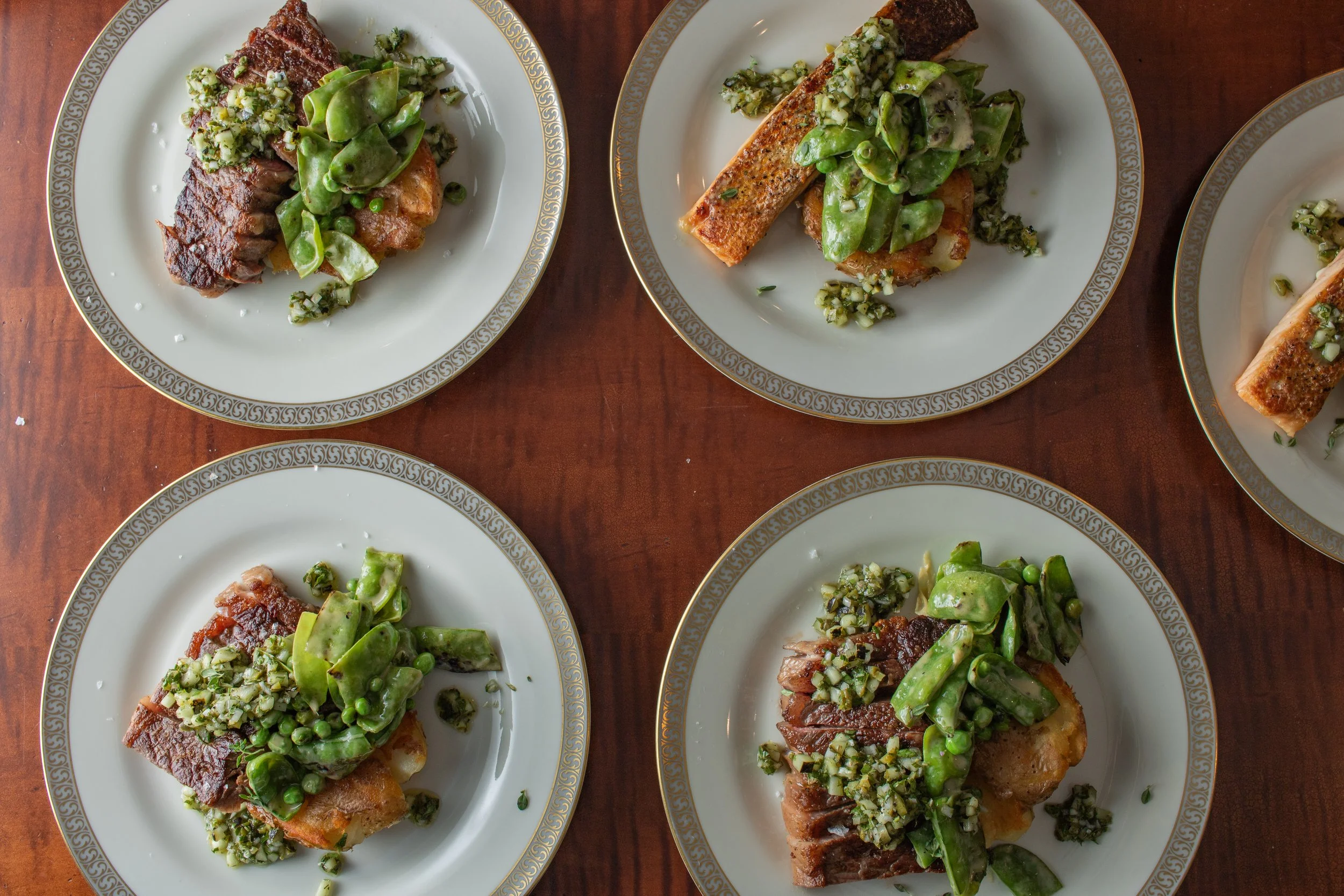 Four plates of food featuring meat topped with green vegetables and garnished with chopped herbs, placed on a wooden table.
