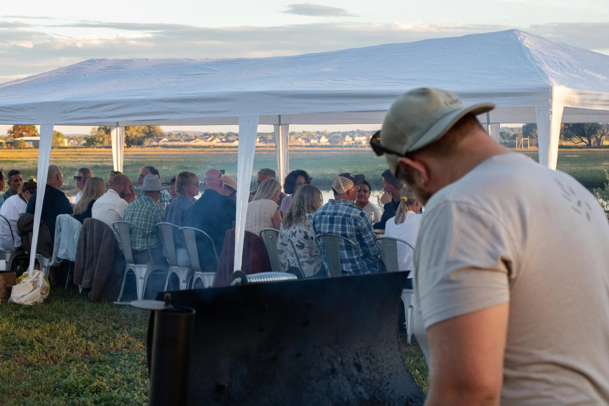 People gathered under a white outdoor tent at an event with a rural landscape in the background, seen in the late afternoon or early evening.