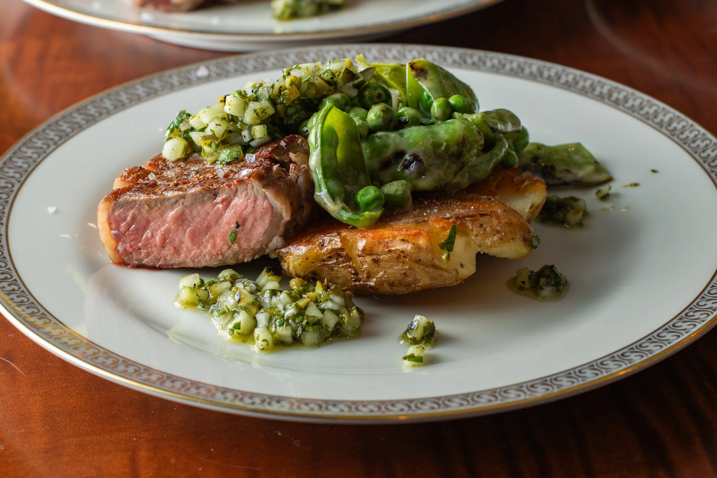 A plated meal with a cut of medium-rare steak topped with a green herb sauce, served with a side of chopped leeks or green onions and peas in a creamy sauce.