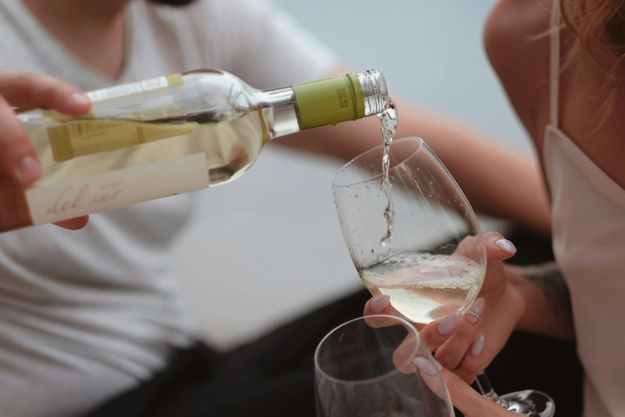Person pouring white wine into a wine glass held by another person.