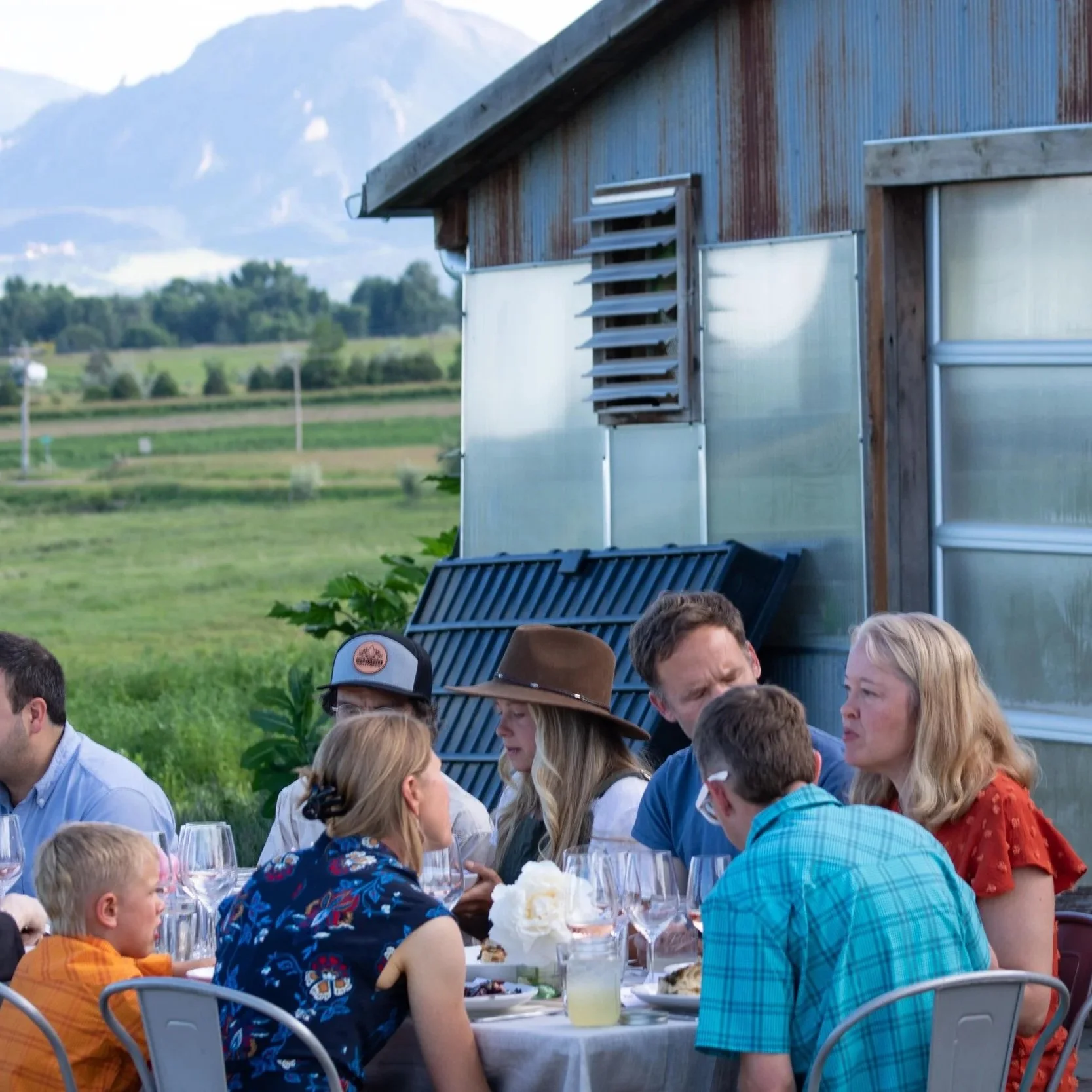 People seated at an outdoor table enjoying a meal, with a rustic background of a weathered barn and green fields in a rural setting.
