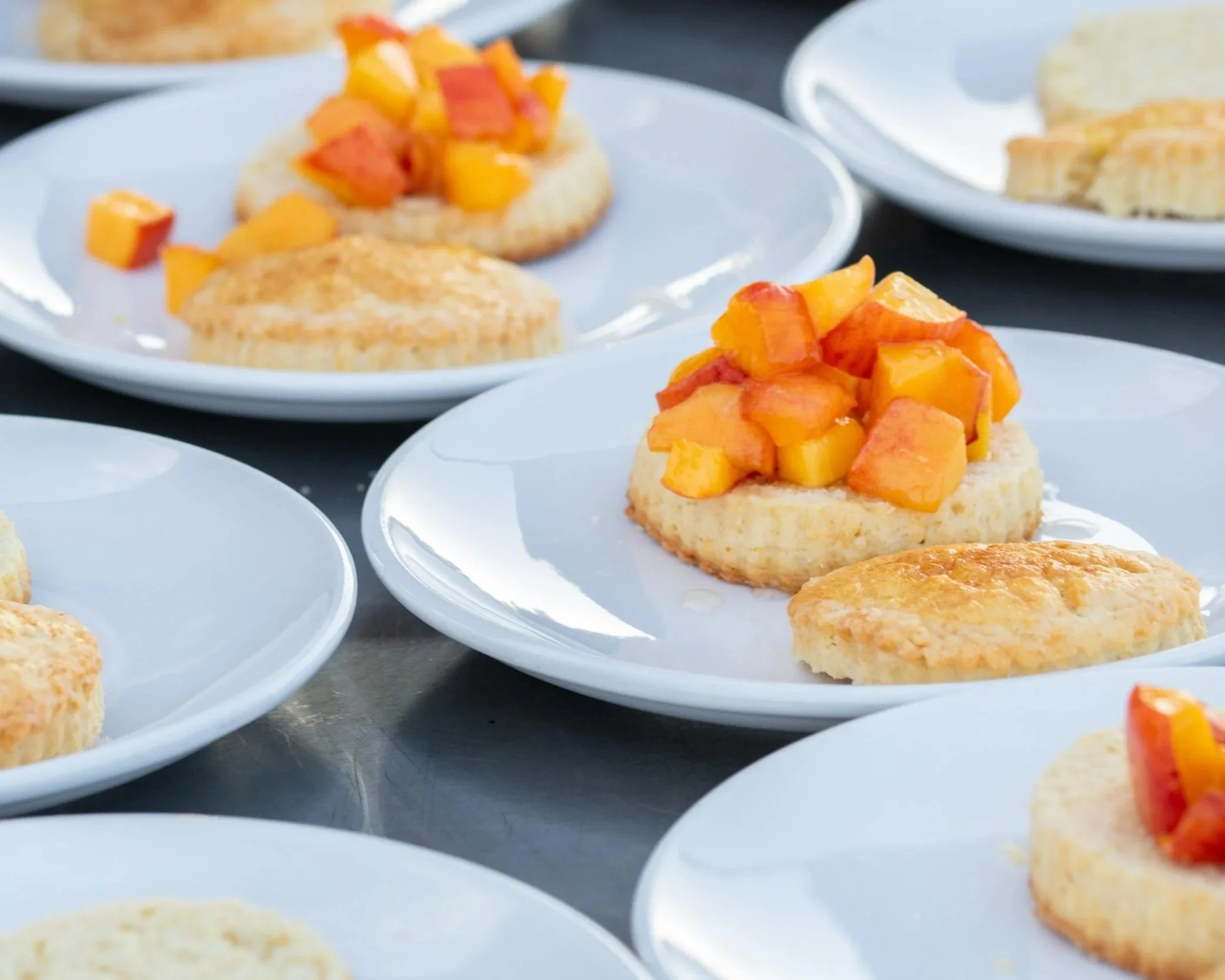 Several white plates with biscuits topped with diced peaches and mangoes on a black surface.
