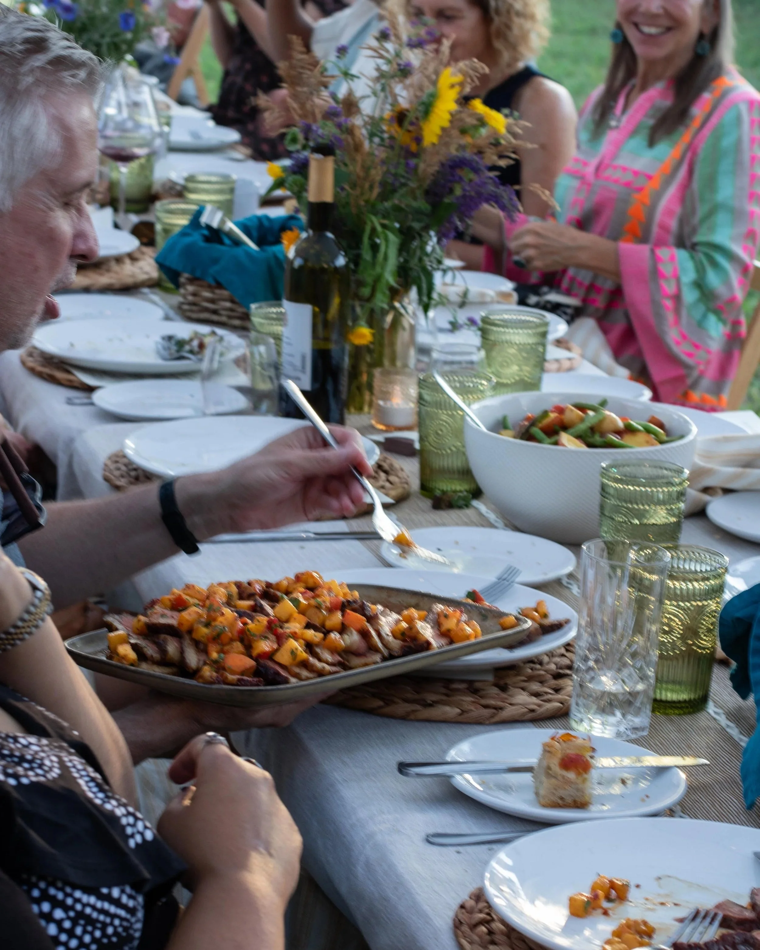 People enjoying an outdoor dinner at a long table with dishes, wine, and a floral centerpiece.