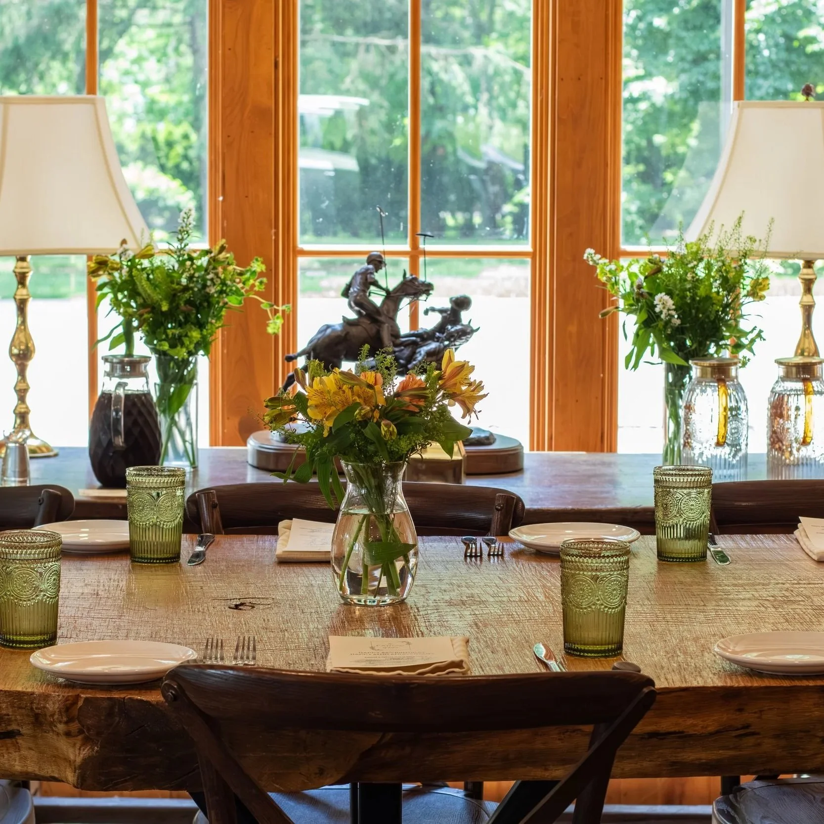 A dining table set with green glasses, white plates, and silverware, decorated with a central glass vase holding yellow lilies and greenery, flanked by two flower arrangements in glass vases, in front of a large window showing outdoor greenery.