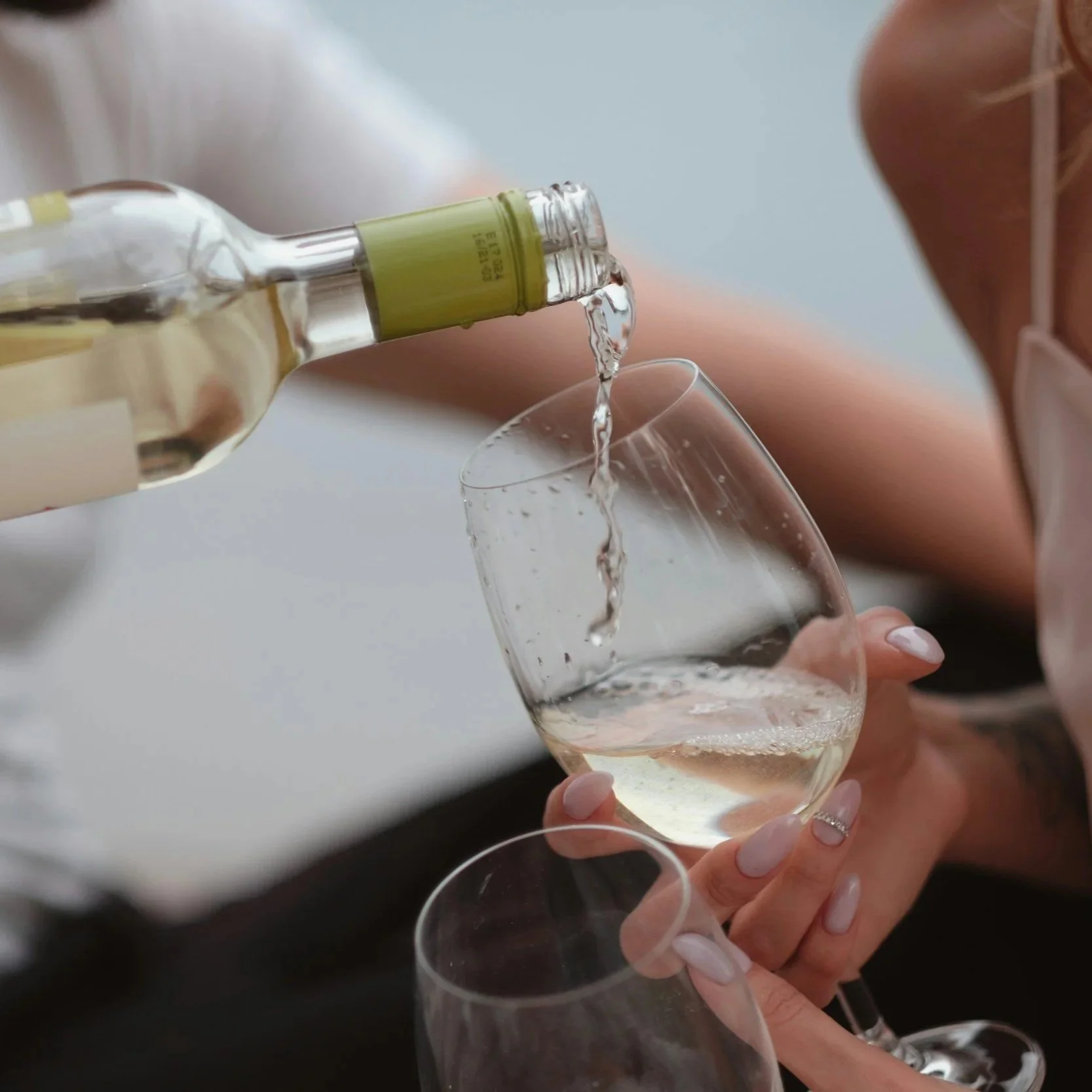 Two women are pouring white wine from a bottle into wine glasses.
