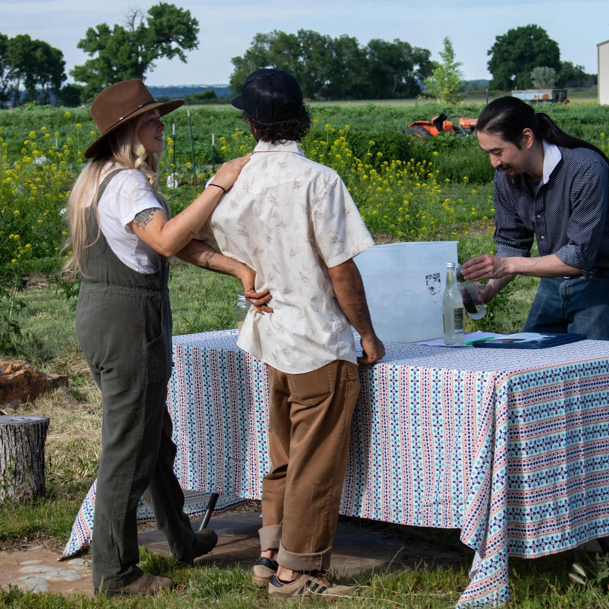 Two women and one man standing outdoors near a table with a colorful cloth, with farm fields and trees in the background, engaging in conversation and laughing.