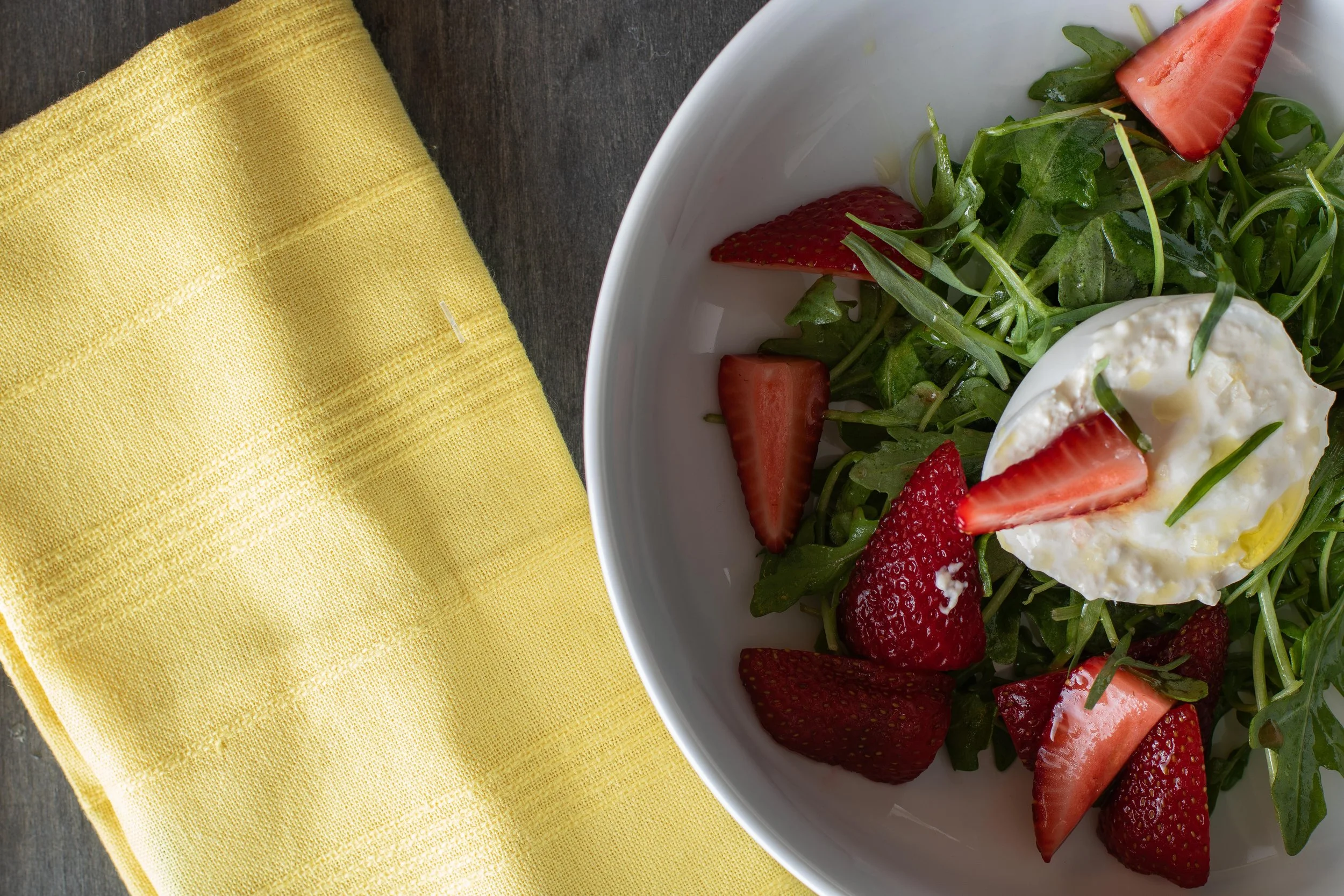 A white bowl containing a salad with sliced strawberries, greens, and a poached egg, placed on a dark surface next to a yellow napkin.