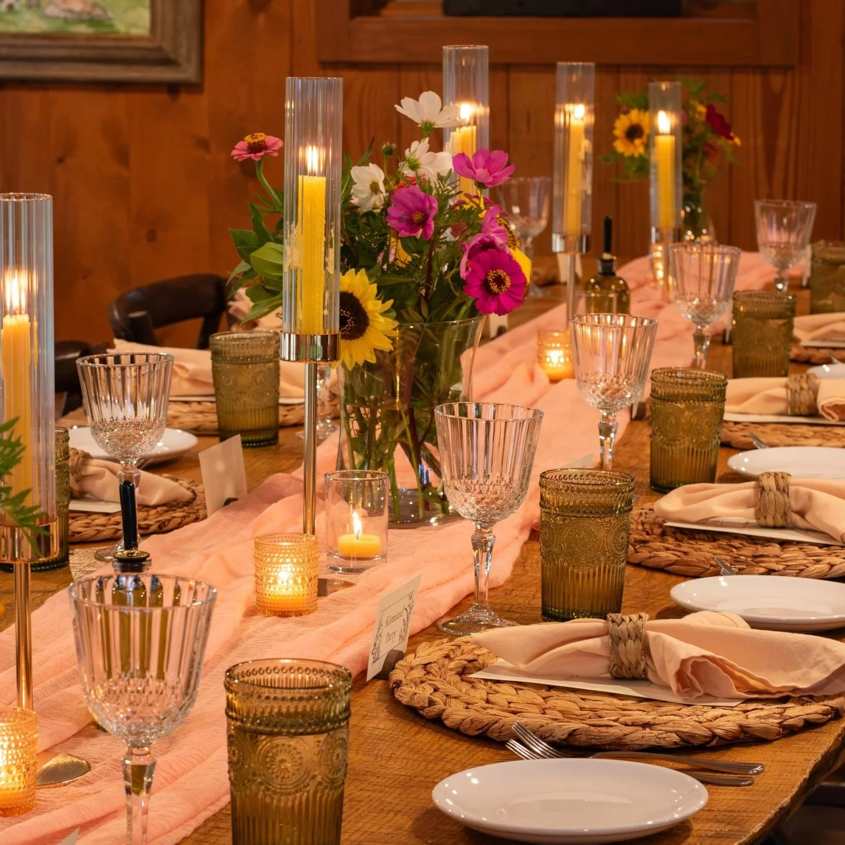 A decorated dining table with floral centerpieces, candles, glassware, plates, and neatly folded napkins in a rustic wood-paneled room.