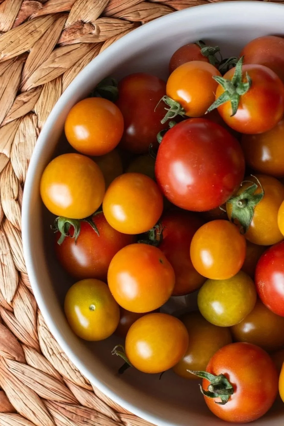 A white bowl filled with various small cherry and heirloom tomatoes in shades of red, yellow, and orange, on a woven straw placemat.